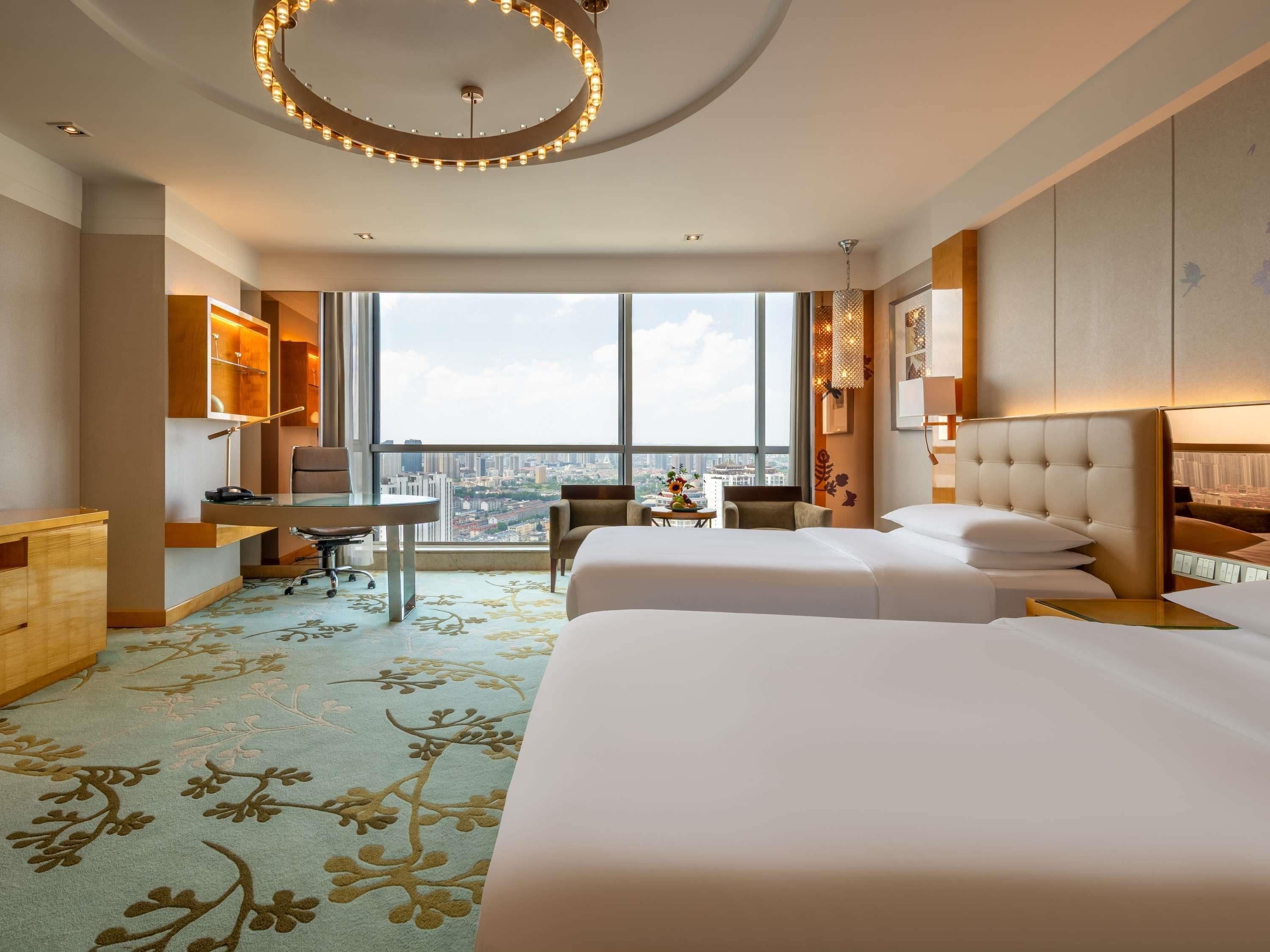 Habitación de hotel con dos camas, un escritorio, una silla, vistas a la ciudad y una lámpara circular grande.