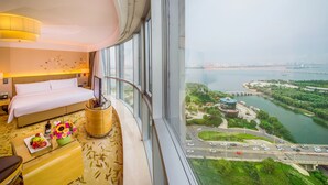 A hotel room with a bed, a glass coffee table with flowers, a TV, and a view of a river and cityscape. - Pullman Linyi Lushang (Linyi)