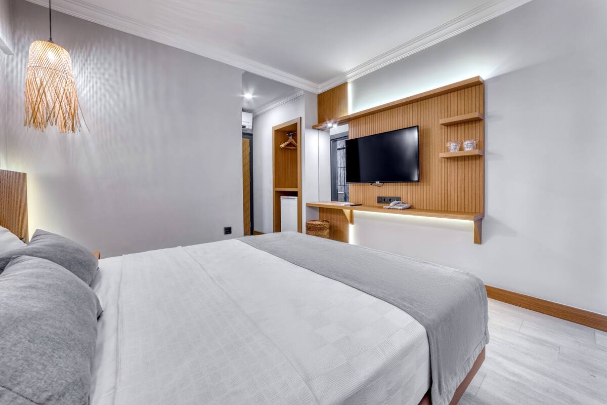 A modern hotel room with a large bed, a TV mounted on a wooden panel wall, and a hanging light fixture.