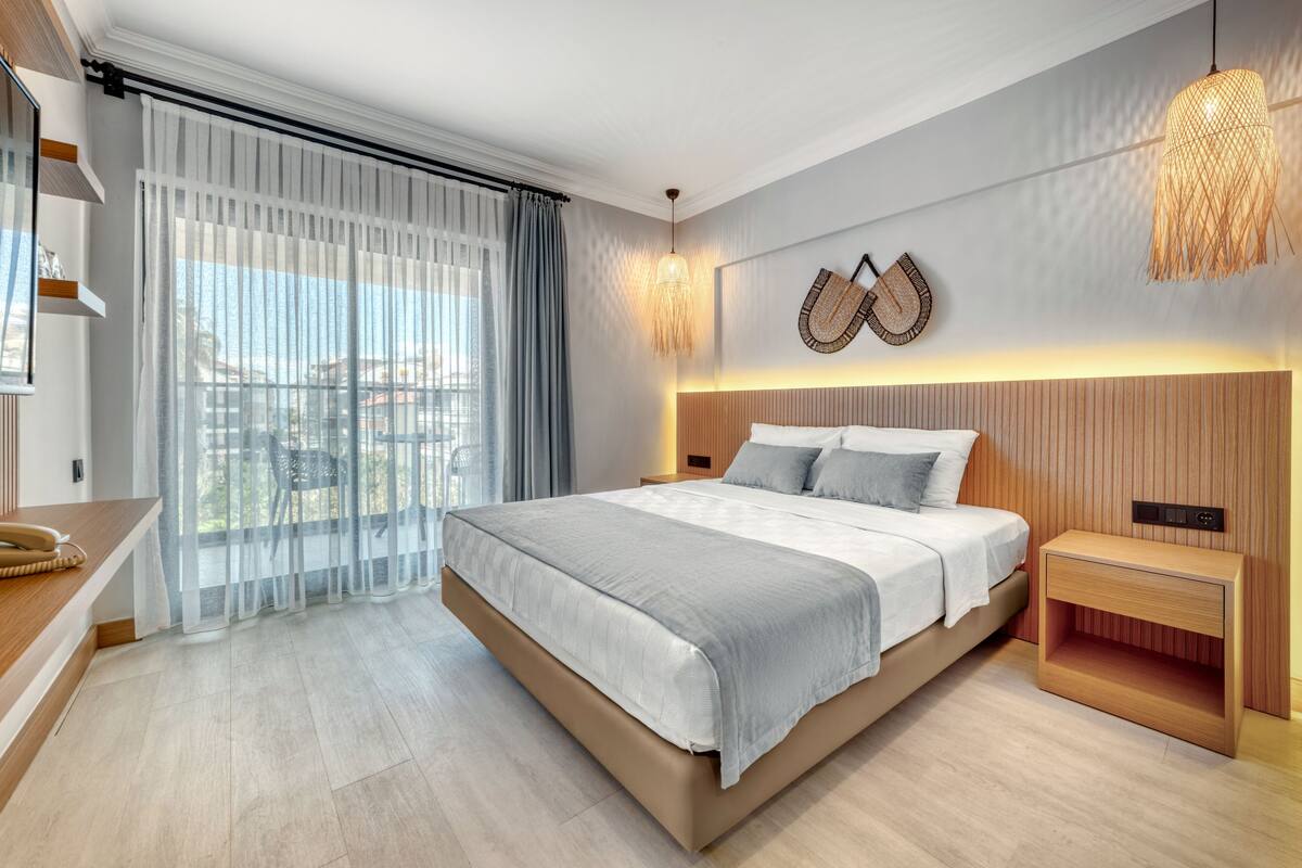 A modern bedroom with a large bed, a wooden headboard, a bedside table, and a view of the city through sheer curtains.