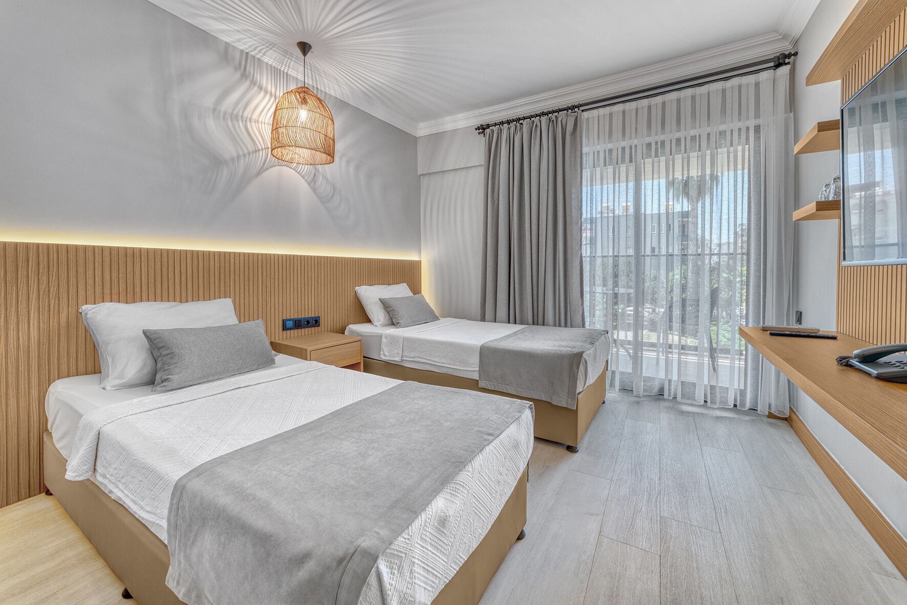Ein modernes Hotelzimmer mit zwei Betten, einem Holzkopfteil, einem Nachttisch, einem Schreibtisch mit Telefon und einem großen Fenster mit transparenten Vorhängen.