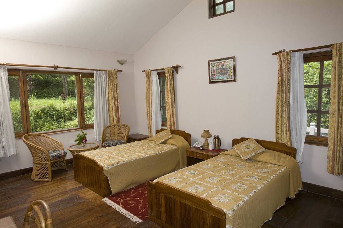 Ein Zimmer mit zwei Holzbetten, einem kleinen Tisch mit Lampe, einem Stuhl und einem Fenster mit Vorhängen.