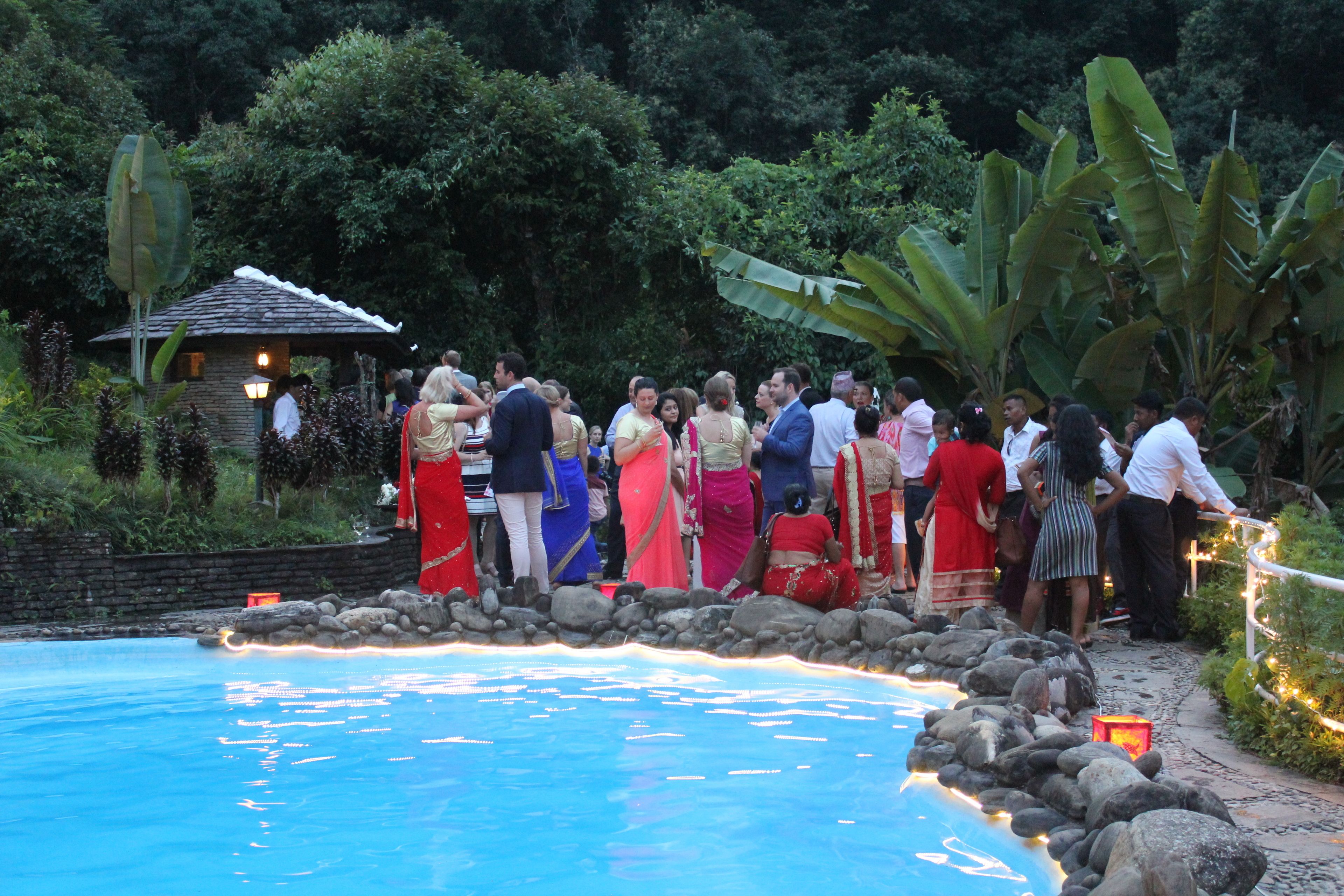 A group of people gathered around a pool with a stone pathway and tropical plants.