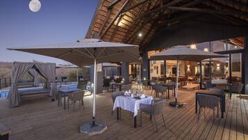 Un espace extérieur aménagé pour les repas, avec des tables et des chaises, une piscine et une structure dotée d’un toit de chaume.