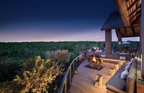 Una terraza de madera con asientos al aire libre, una fogata y vistas a un paisaje boscoso bajo un cielo estrellado.