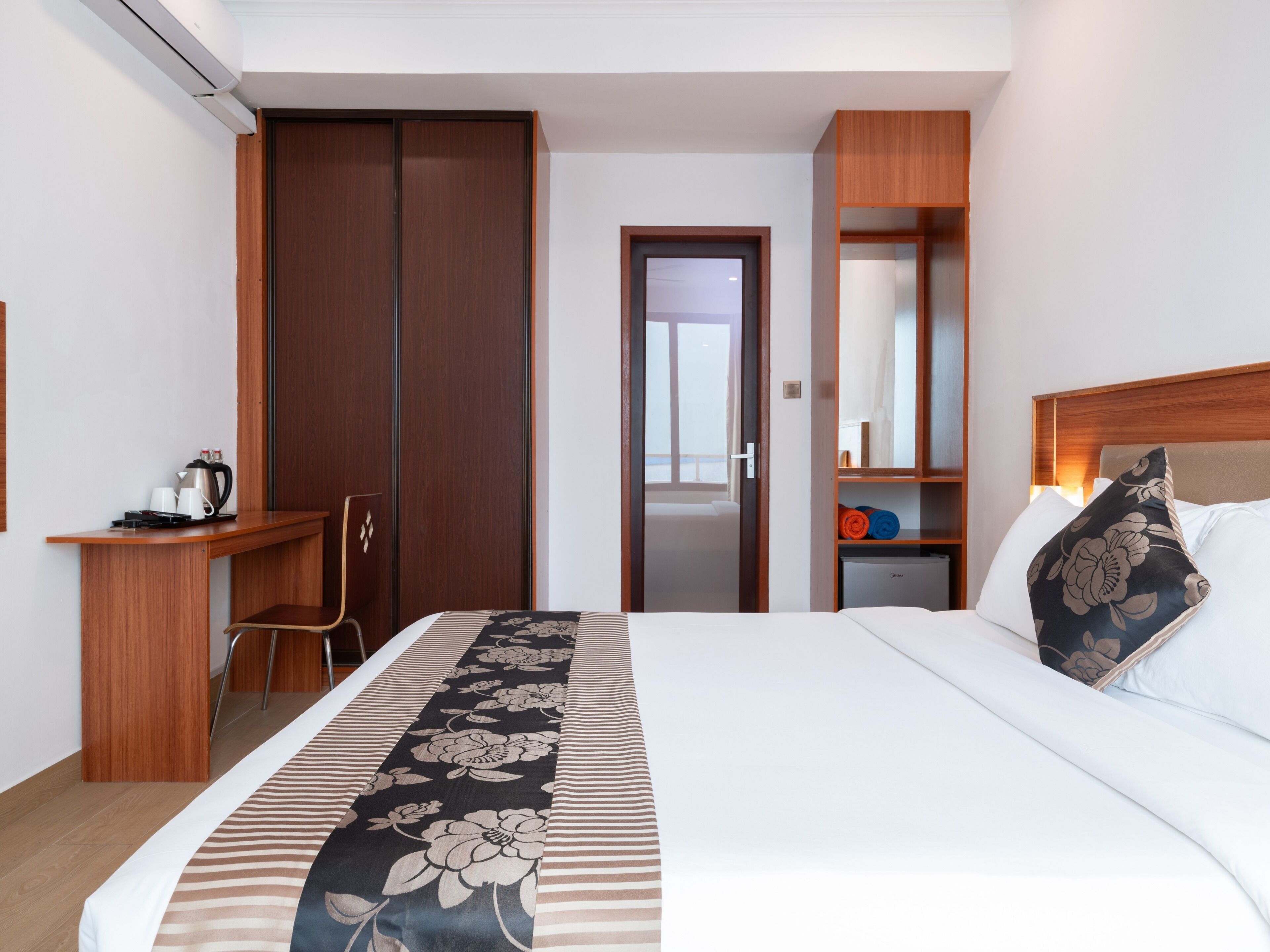 A hotel room with a bed, a desk, a chair, a wardrobe, a nightstand, and a mirror.