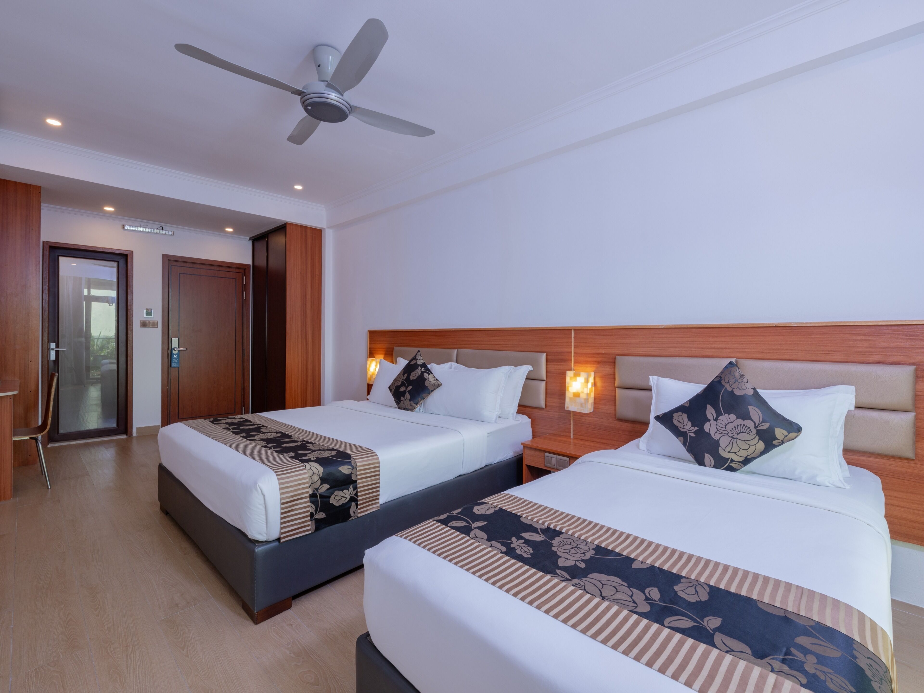 Una habitación de hotel con dos camas, un ventilador de techo, un cabecero de madera y una mesita de noche con lámpara.