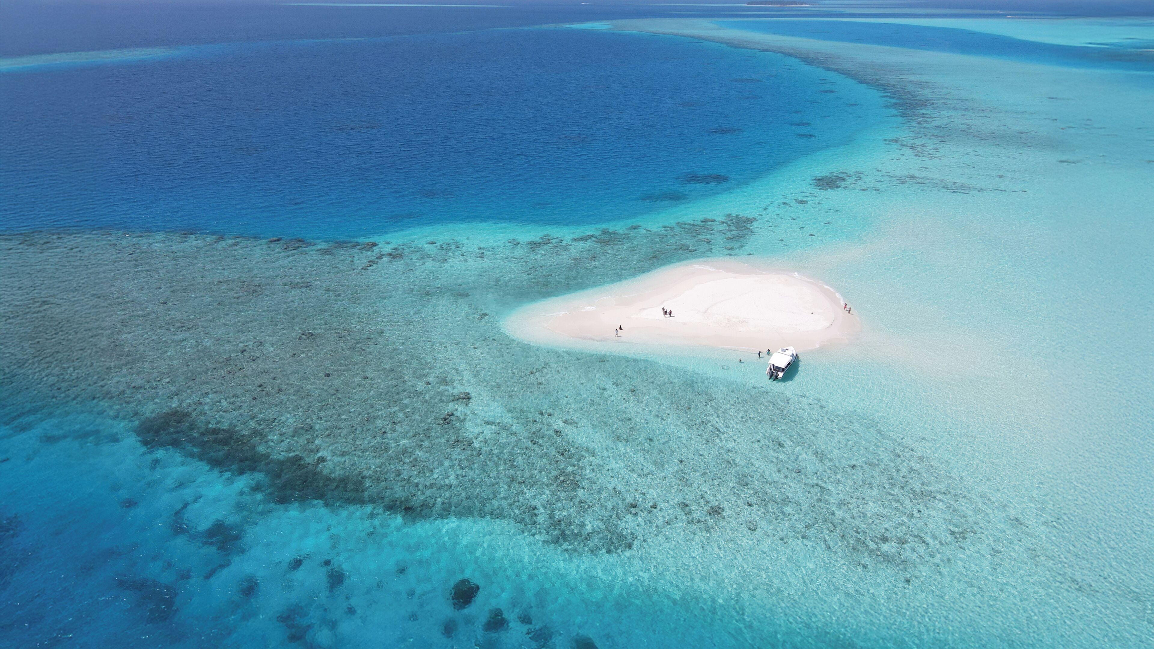 An aerial view of a beach with clear turquoise waters, a sandy island, and a small boat.