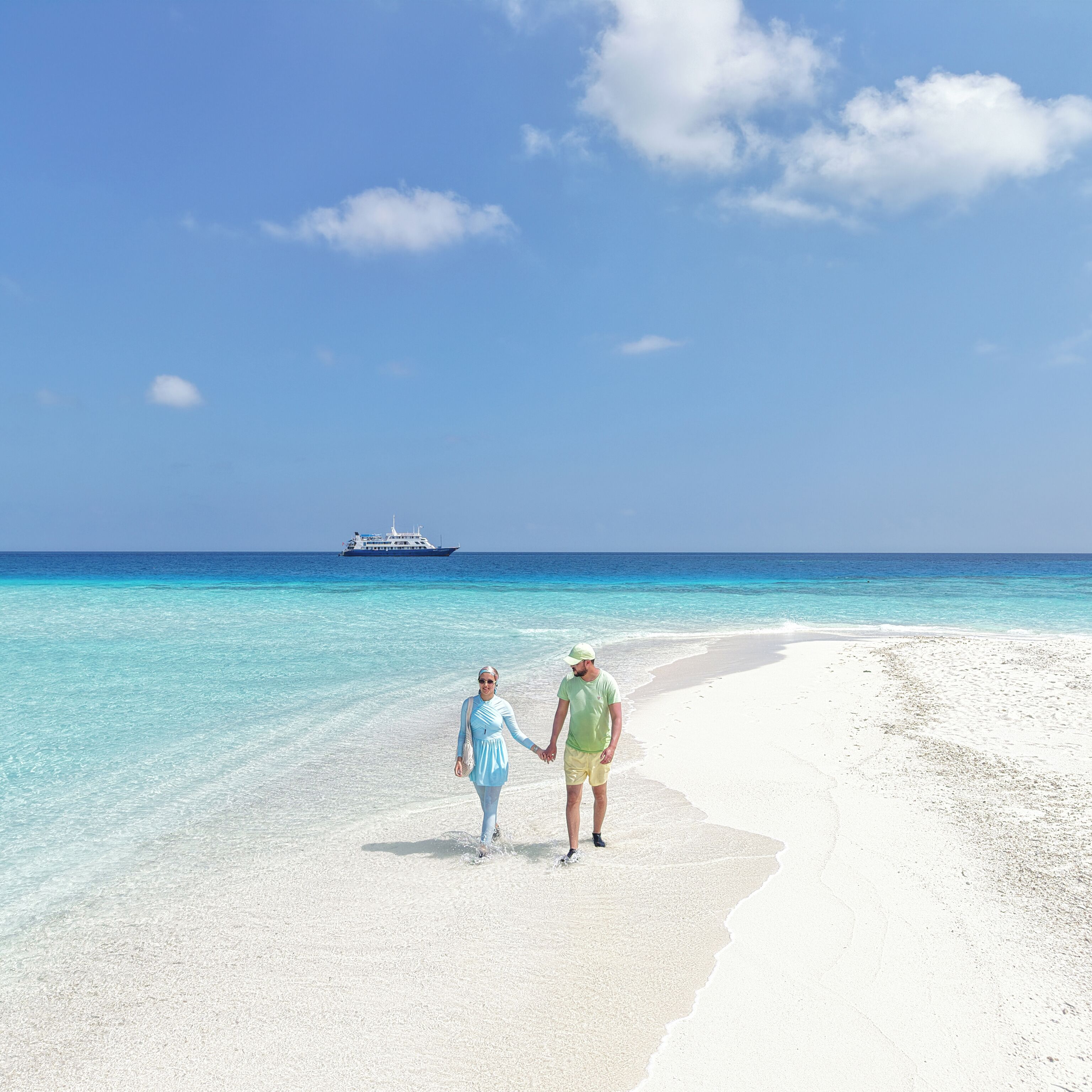 A beach with clear turquoise water, a white sandy shore, and a ship in the distance.