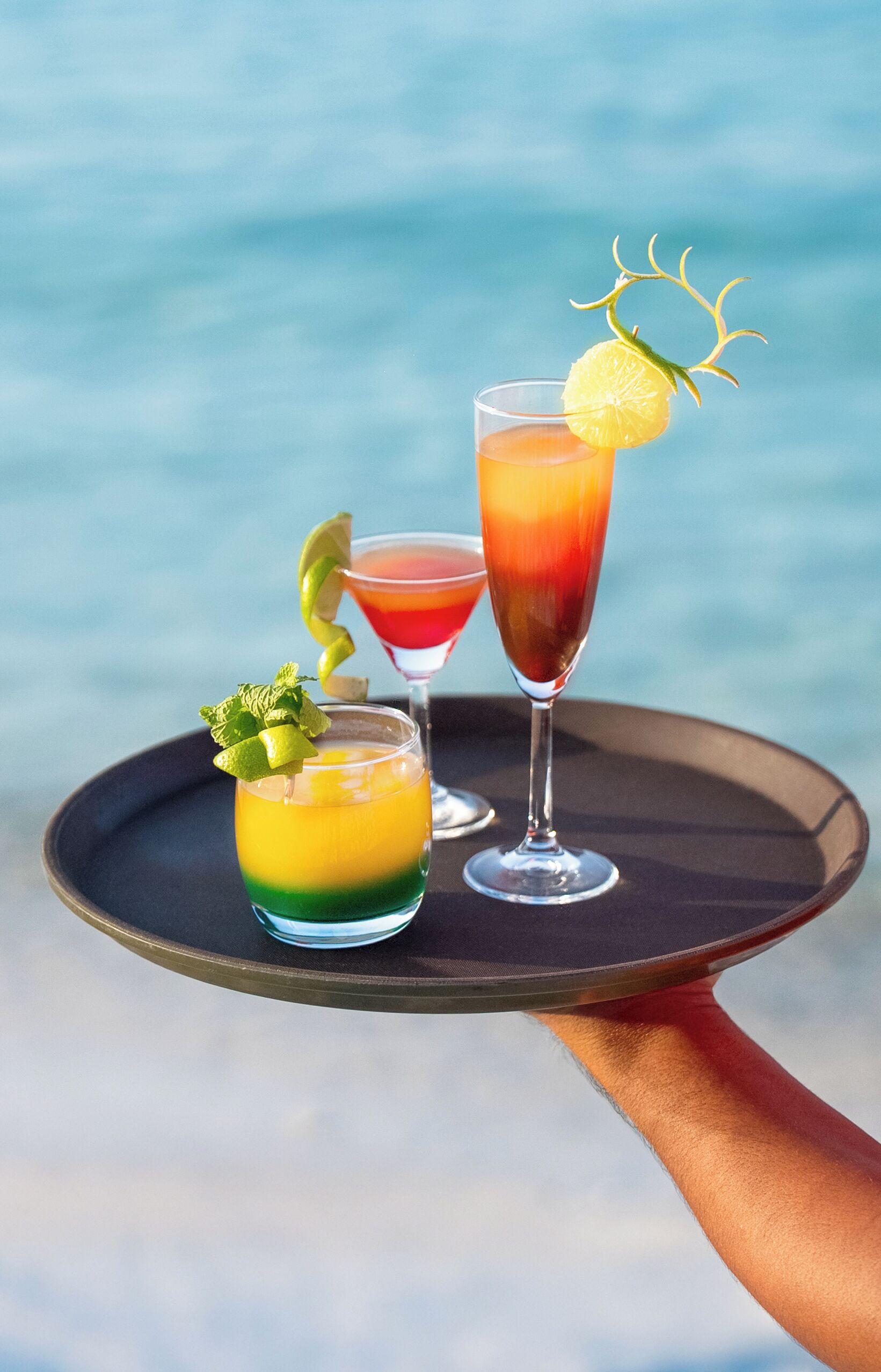 Three cocktails on a tray with a blurred ocean background.