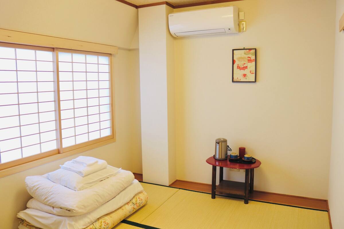 A room with tatami flooring, a bed with neatly folded white linens, a small table with a kettle and cups, and a wall-mounted air conditioner.