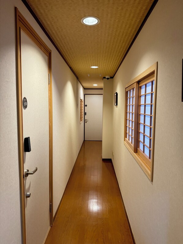 Un pasillo con piso de madera, una puerta con placa de número y una ventana con diseño de cuadrícula.