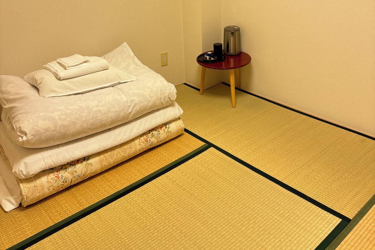 A traditional Japanese-style room with tatami flooring, a bed with neatly folded white bedding, a small table with a red base, and a window with wooden frames.
