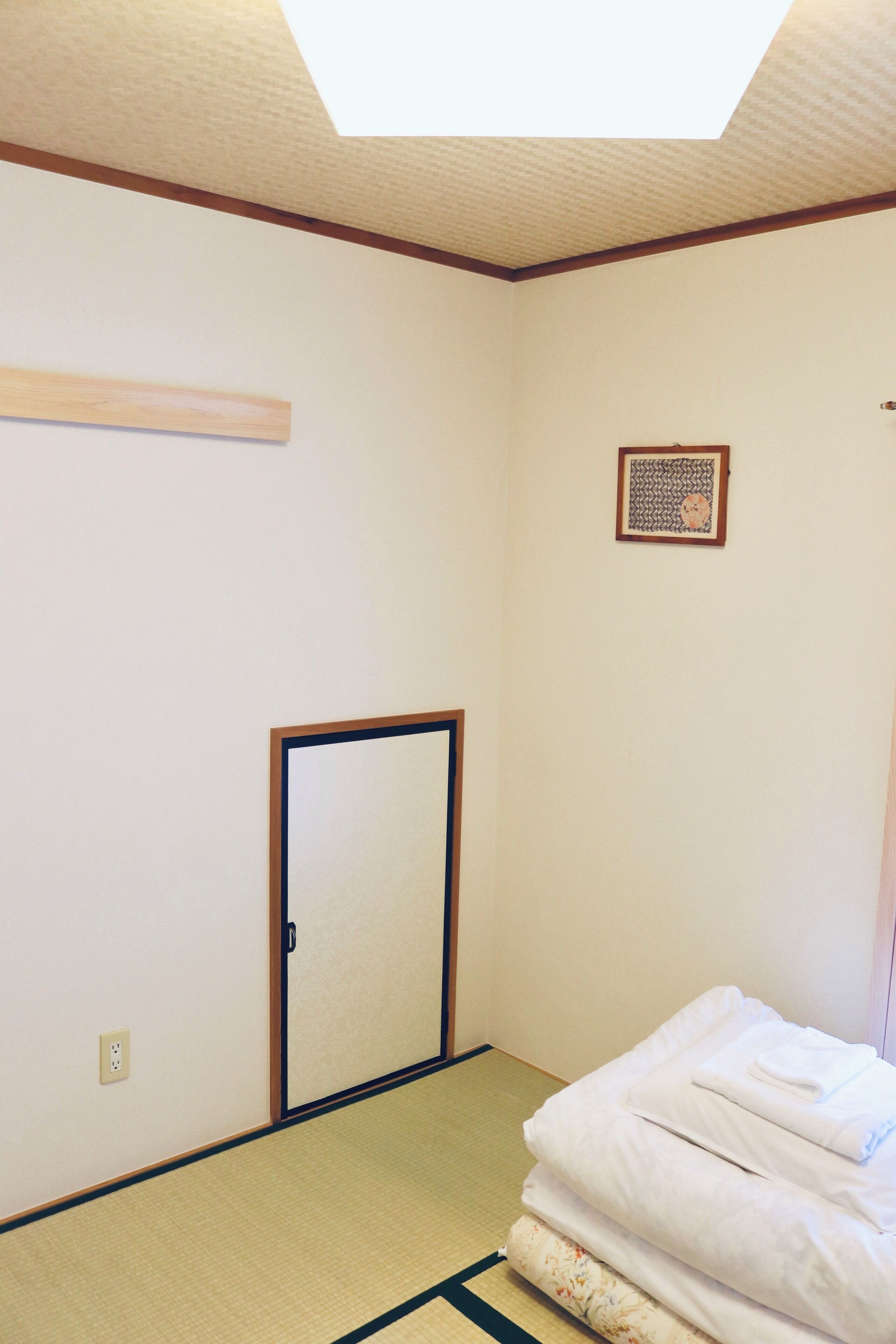 Una habitación con suelo de tatami, puerta corredera shoji, una pequeña obra de arte enmarco y una cama bien hecha con ropa blanca doblada.