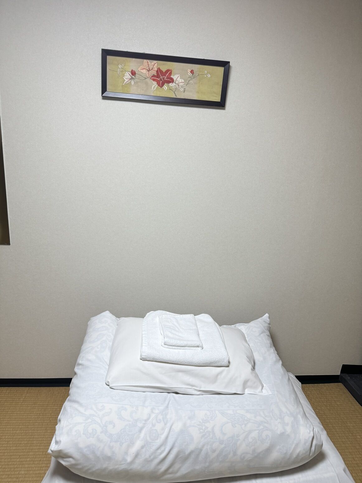 A neatly made bed with a folded towel, a framed floral artwork on the wall, and a plain wall.