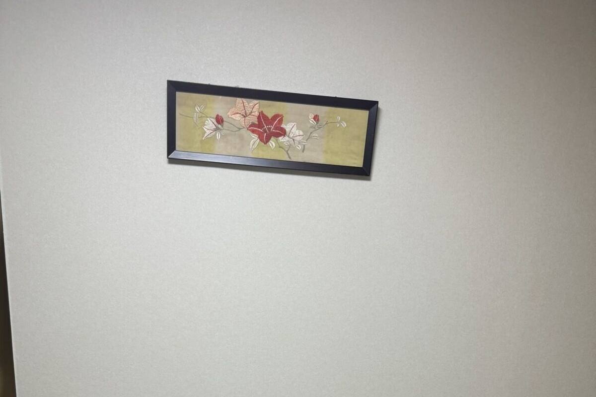 A neatly made bed with a folded towel, a framed floral artwork on the wall, and a plain wall.