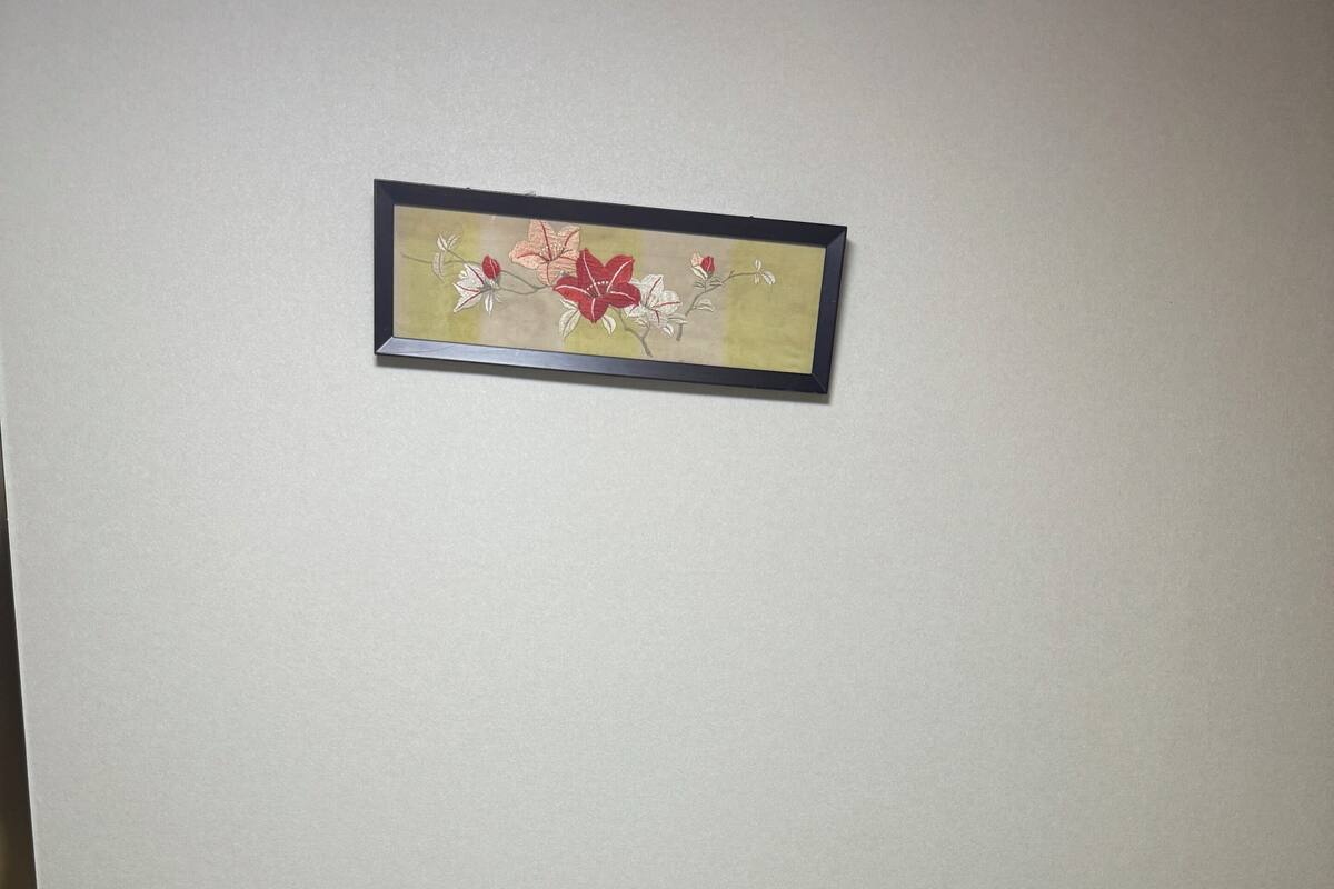 A neatly made bed with a folded white towel and a decorative pillow, a framed floral artwork on the wall, and a plain beige wall.