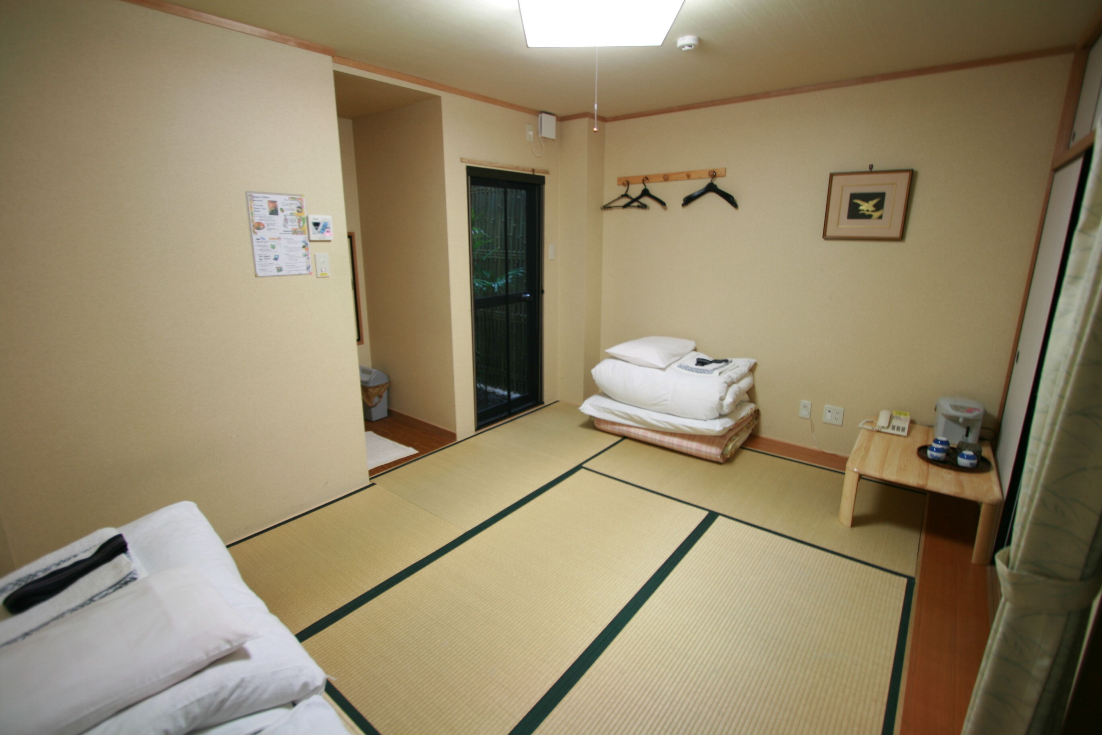A hotel room with tatami flooring, a bed with white bedding, a small table with a kettle, and a wall-mounted coat rack.