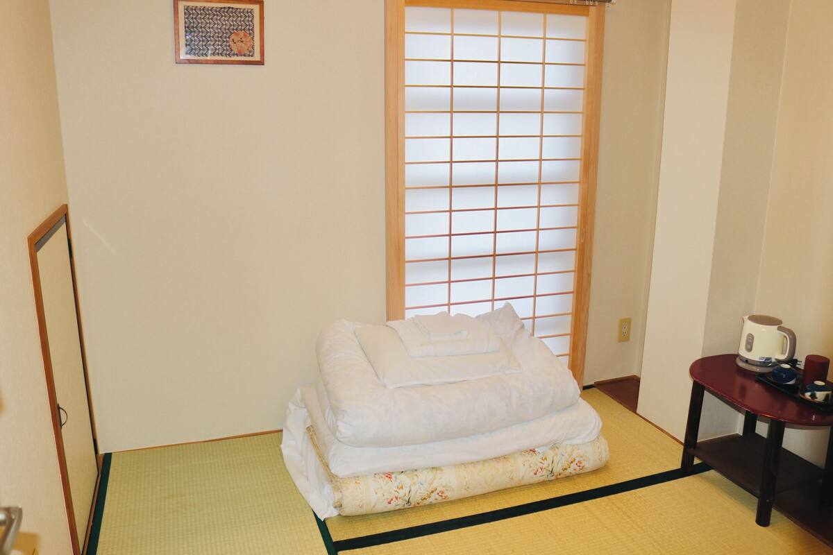 A room with tatami flooring, a bed with white bedding, a shoji screen, a small table with a kettle and cup, and a framed picture on the wall.