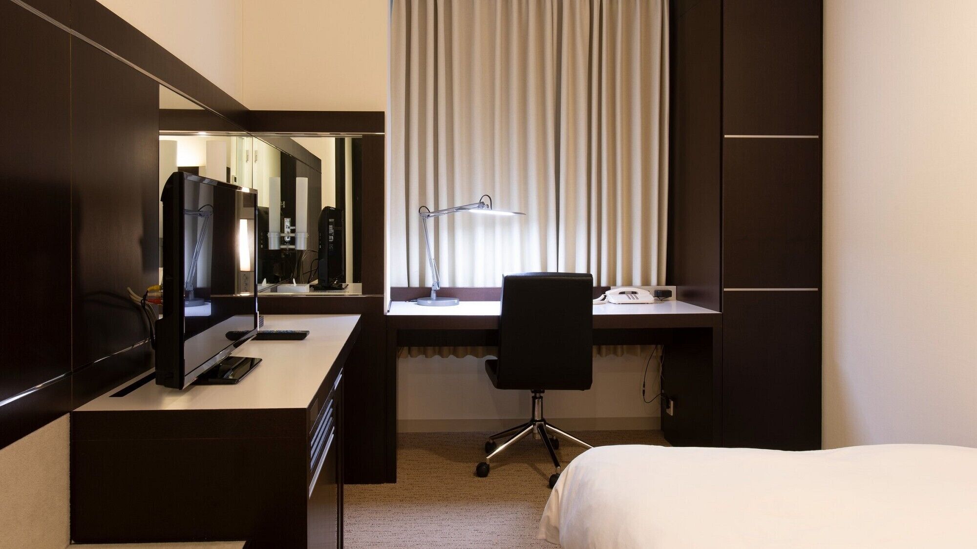 A hotel room with a bed, a desk with a computer, a chair, a telephone, and a window with blinds.