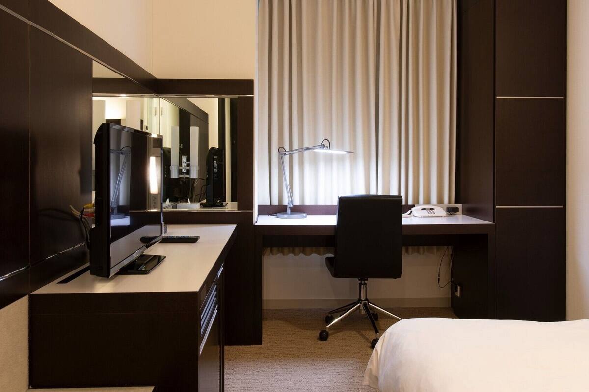 A hotel room with a bed, a desk with a computer, a chair, a telephone, and a window with blinds.