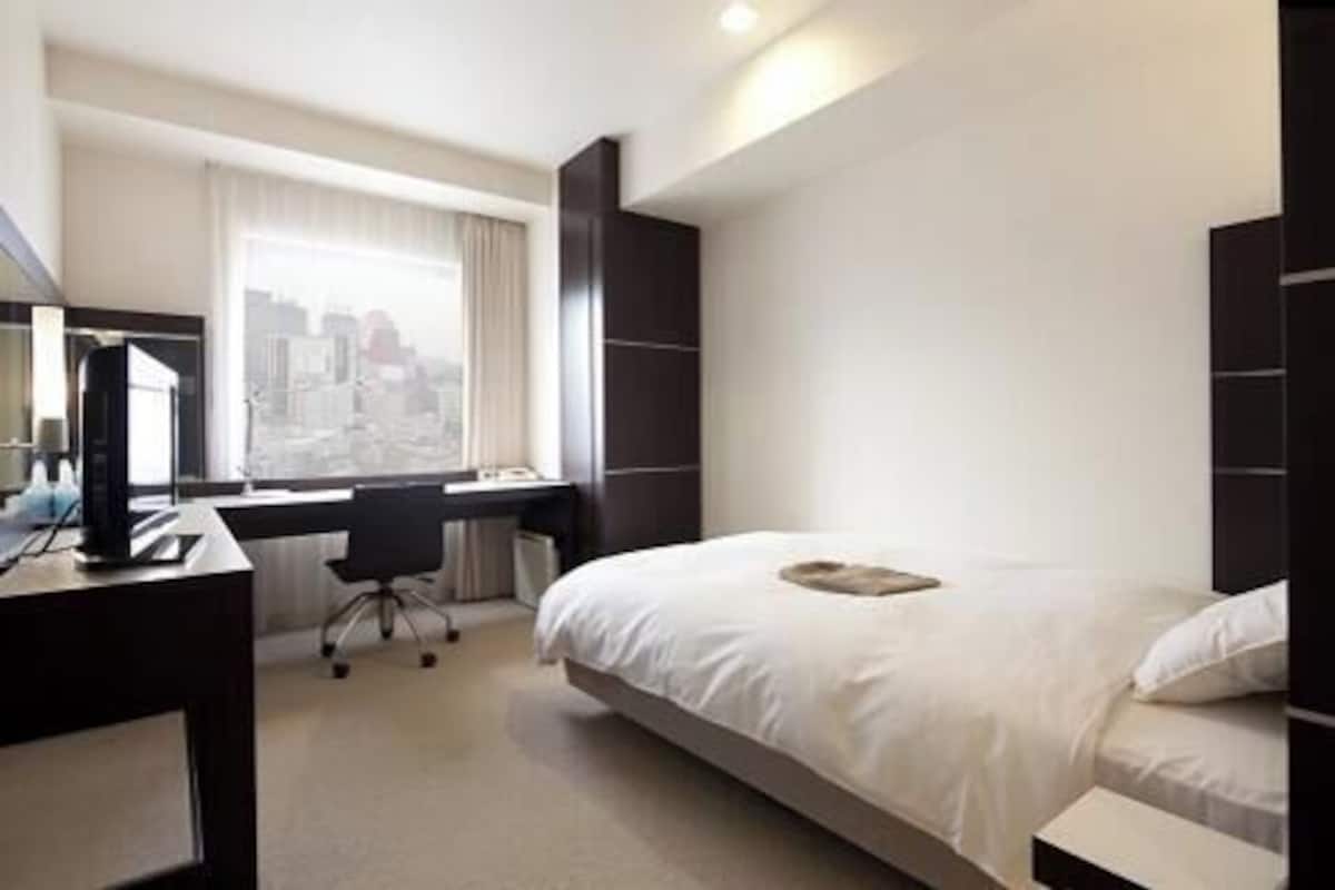 A hotel room with a bed, a desk, a chair, and a large window.