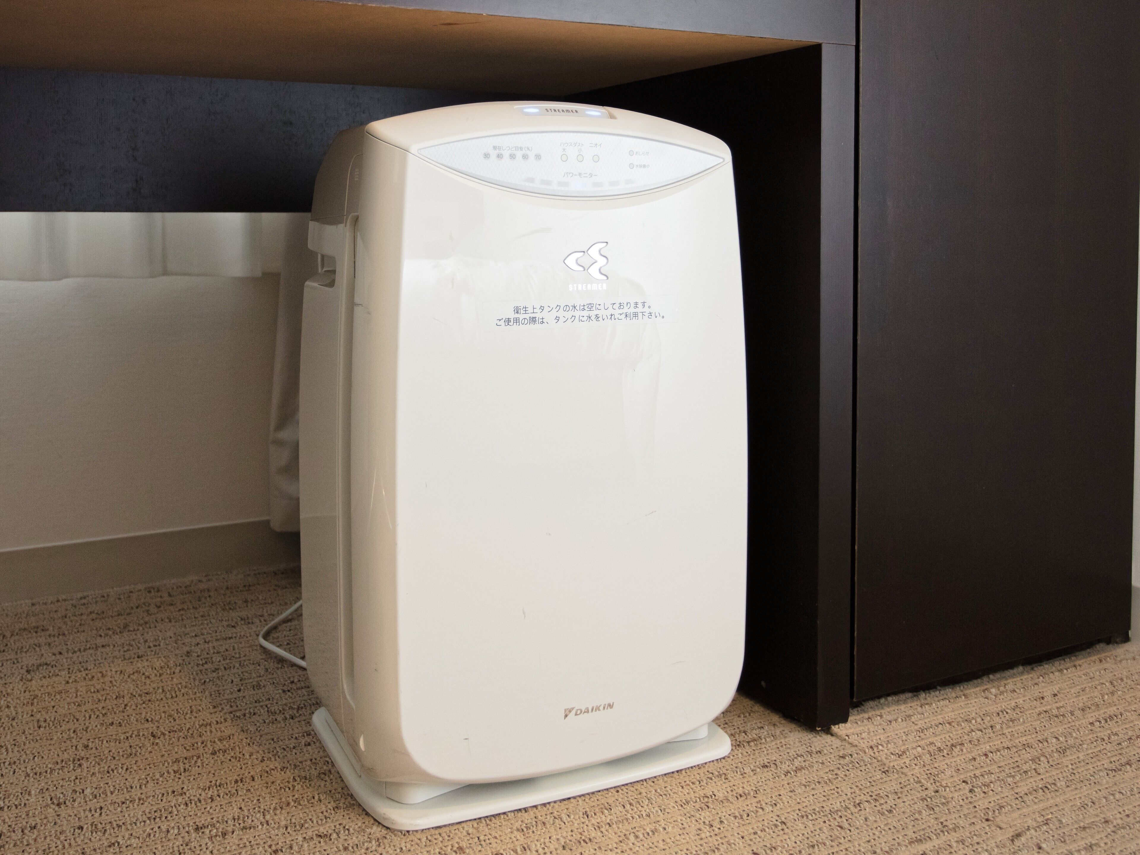 A white Daikin air purifier with a control panel, placed on a carpeted floor under a dark cabinet.