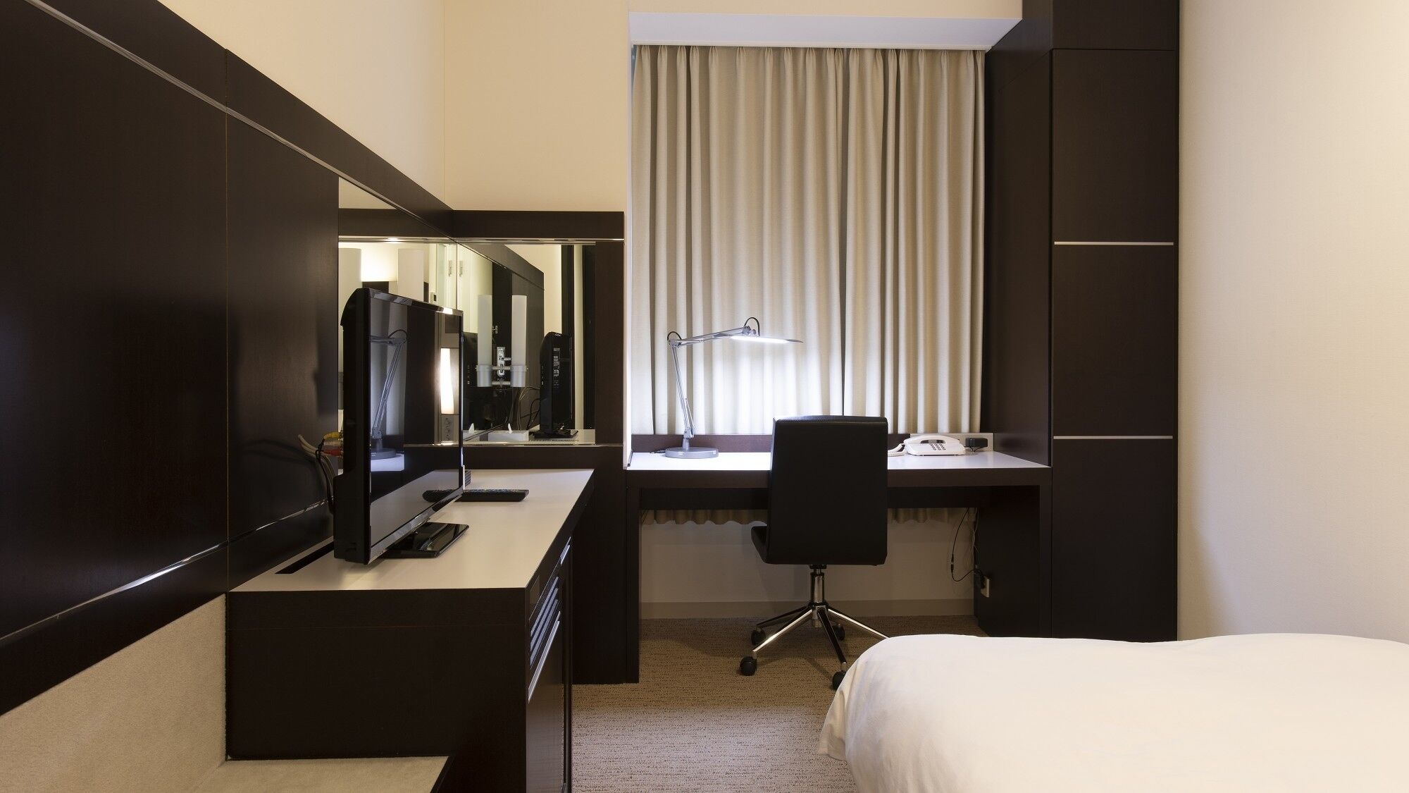 A hotel room with a bed, a desk with a computer and phone, a chair, a desk lamp, and a large mirror.
