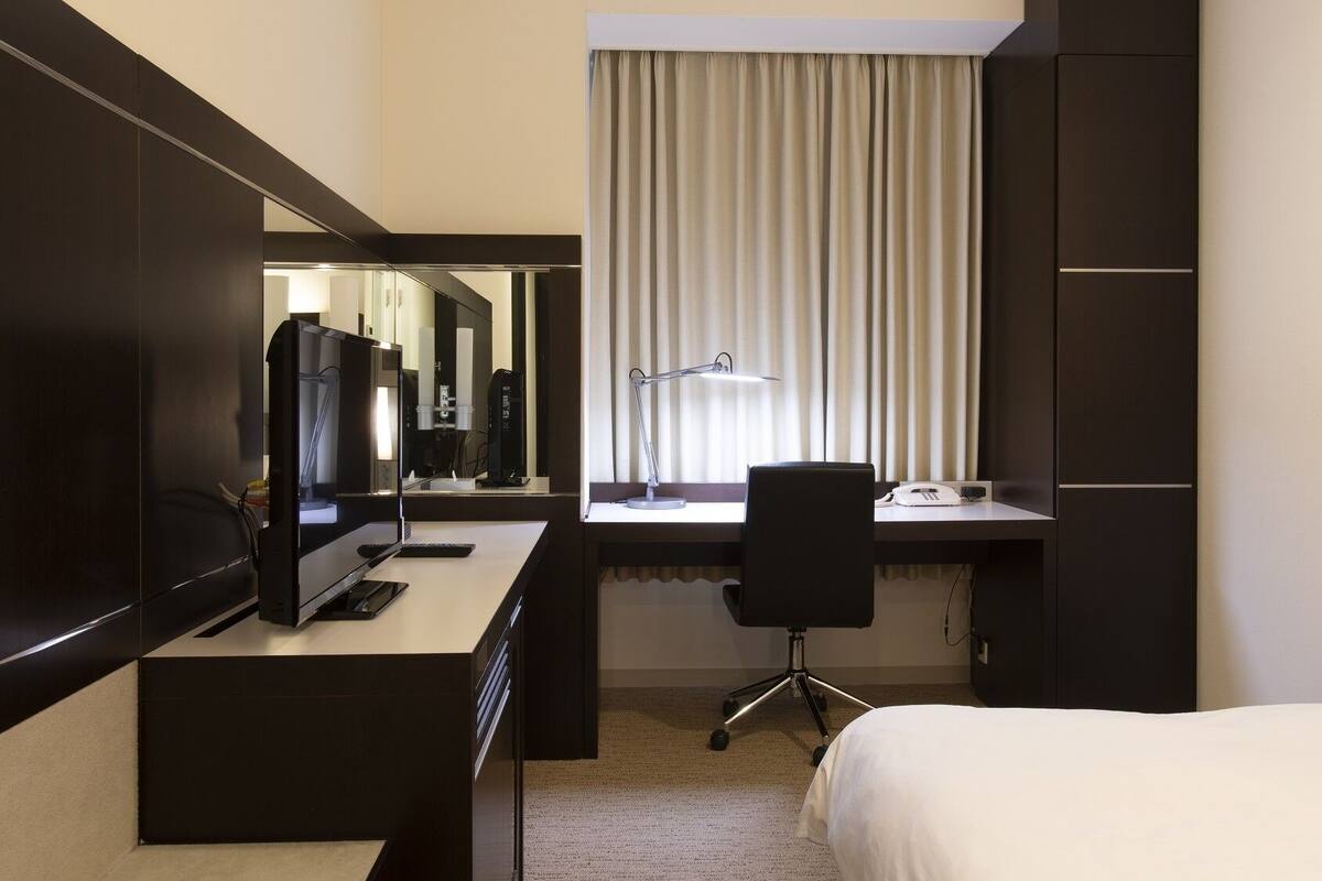 A hotel room with a bed, a desk with a computer and phone, a chair, a desk lamp, and a large mirror.