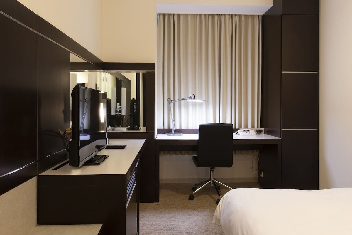 A hotel room with a bed, a desk with a computer and phone, a chair, a desk lamp, and a large mirror.