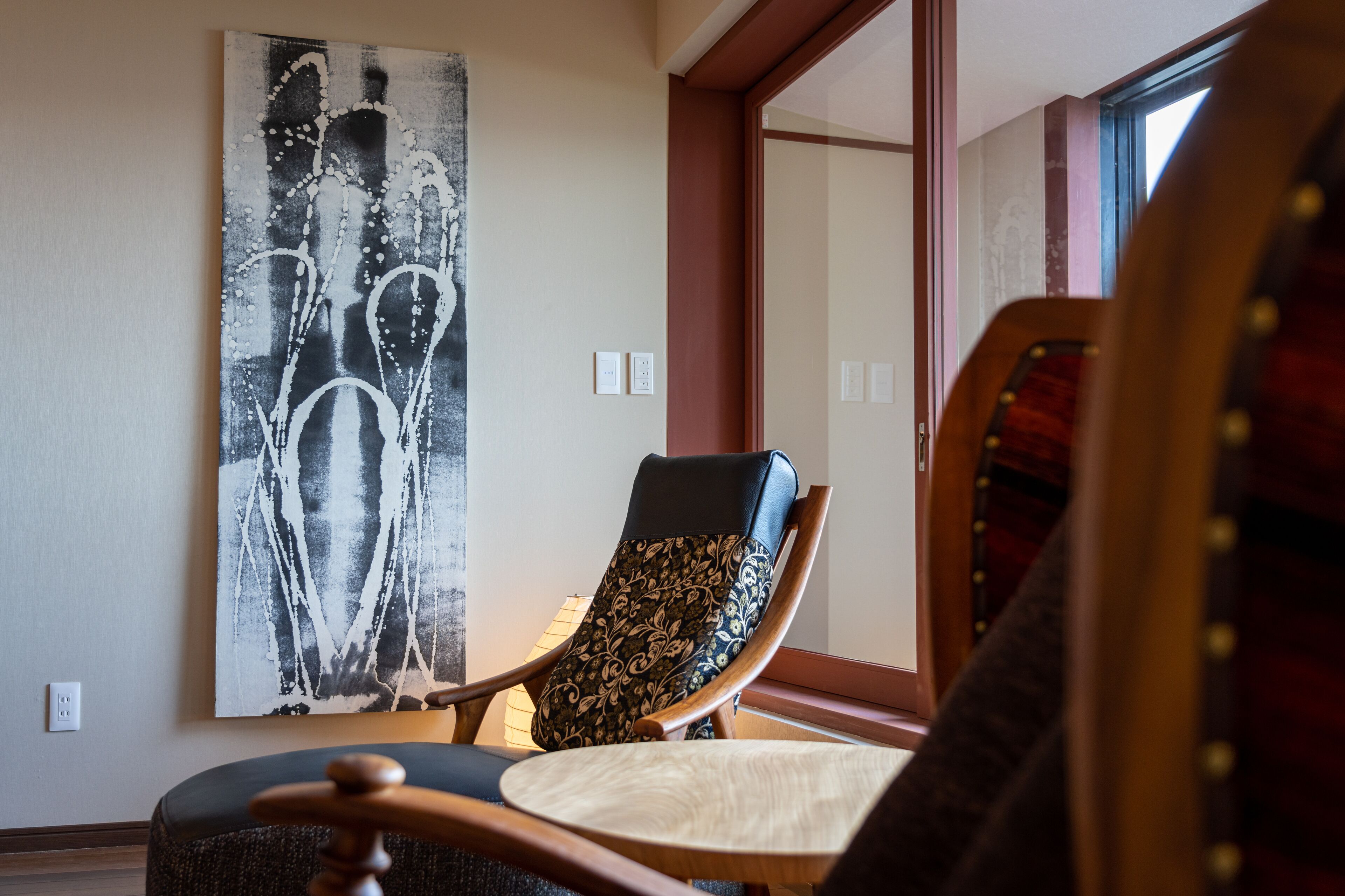 Une pièce avec un grand tableau abstrait au mur, une chaise en bois avec un coussin à motifs et une fenêtre donnant sur l’extérieur.