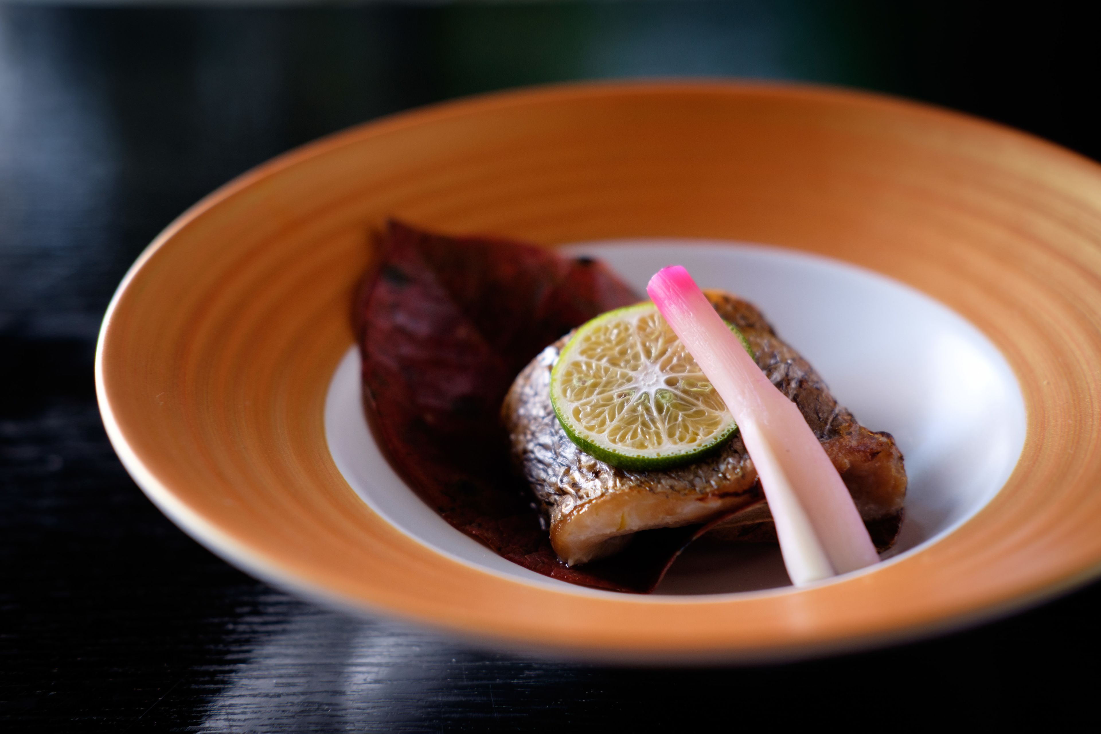 Une assiette contenant un morceau de poisson, une tranche de citron vert et une garniture rose, le tout posé sur une surface sombre.