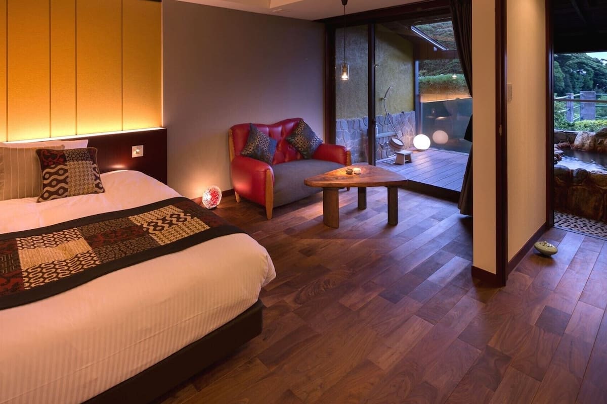 A bedroom with a large bed, a red chair, a small table, and a view of a garden through a glass door.