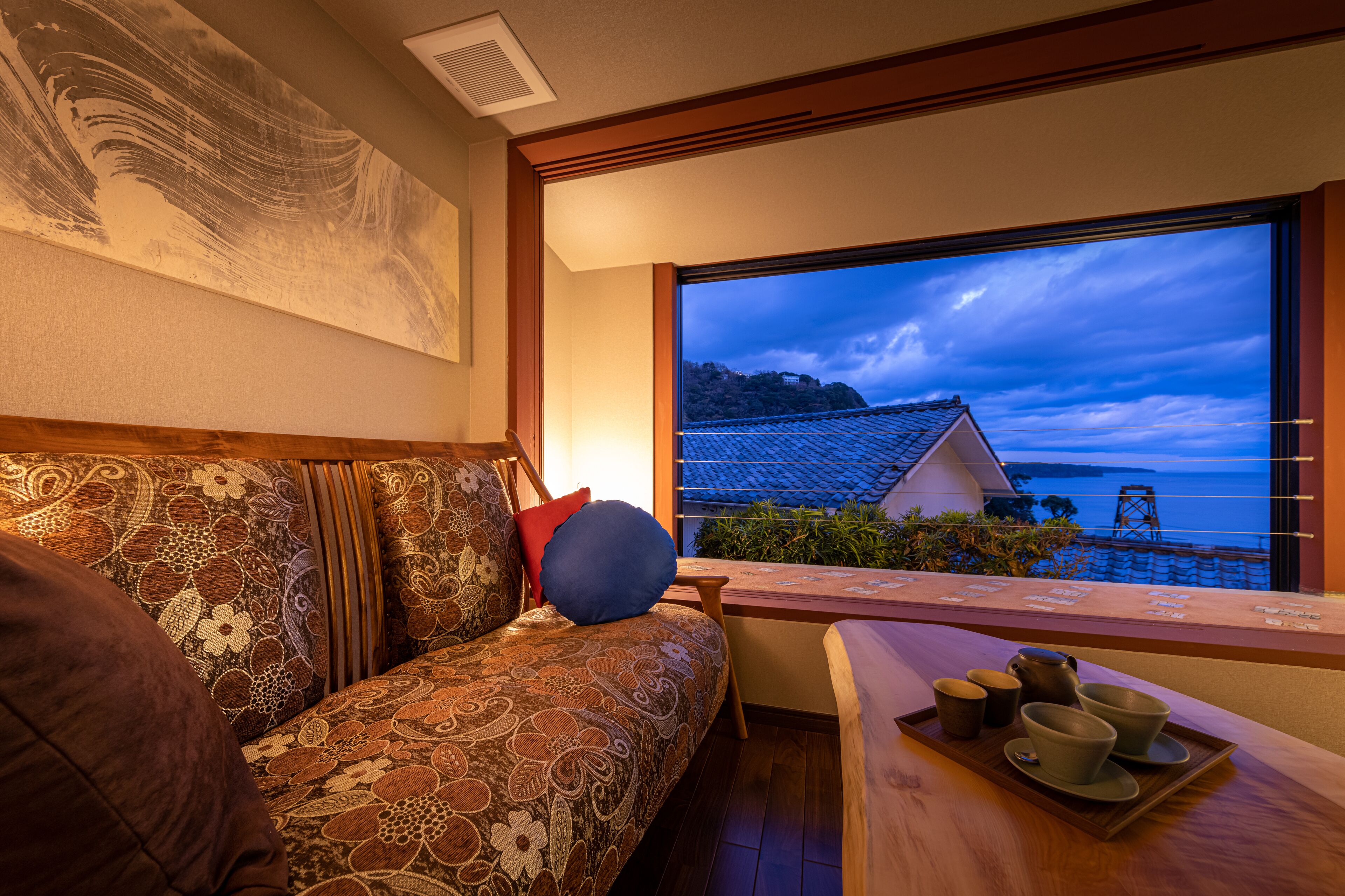 A room with a sofa, a wooden table with cups and a tray, and a view of a house by the sea.