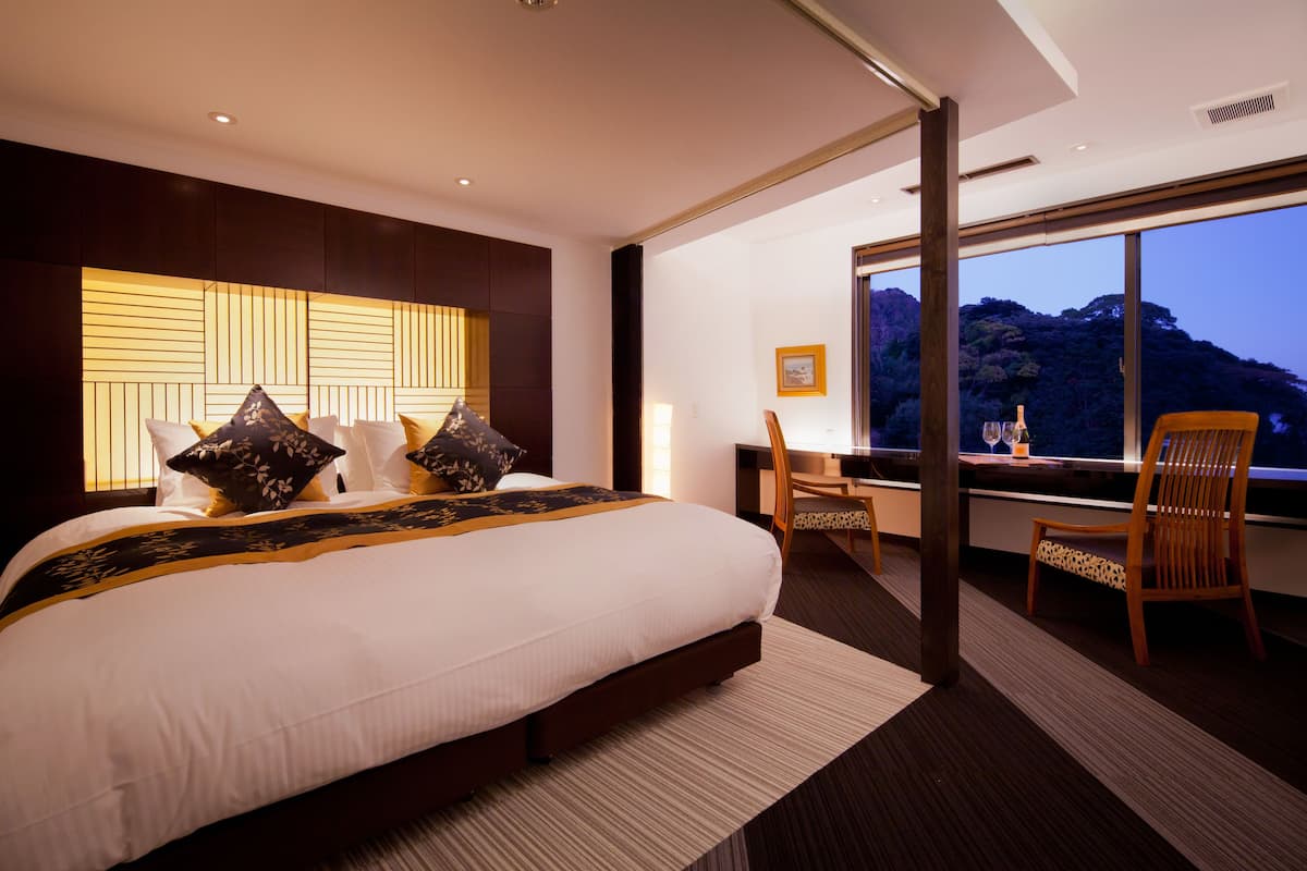 A modern hotel room with a large bed, a desk, and a view of a mountain at dusk.