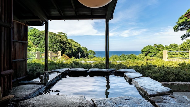 A traditional Japanese onsen with a stone bath surrounded by lush greenery and a view of the sea. - Isaribi (Higashiizu)