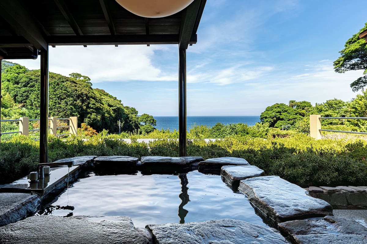 A traditional Japanese onsen with a stone bath surrounded by lush greenery and a view of the sea.