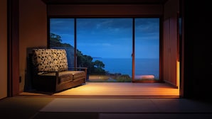 A room with a sofa facing a large sliding glass door leading to a balcony with a view of the sea. - Isaribi (Higashiizu)