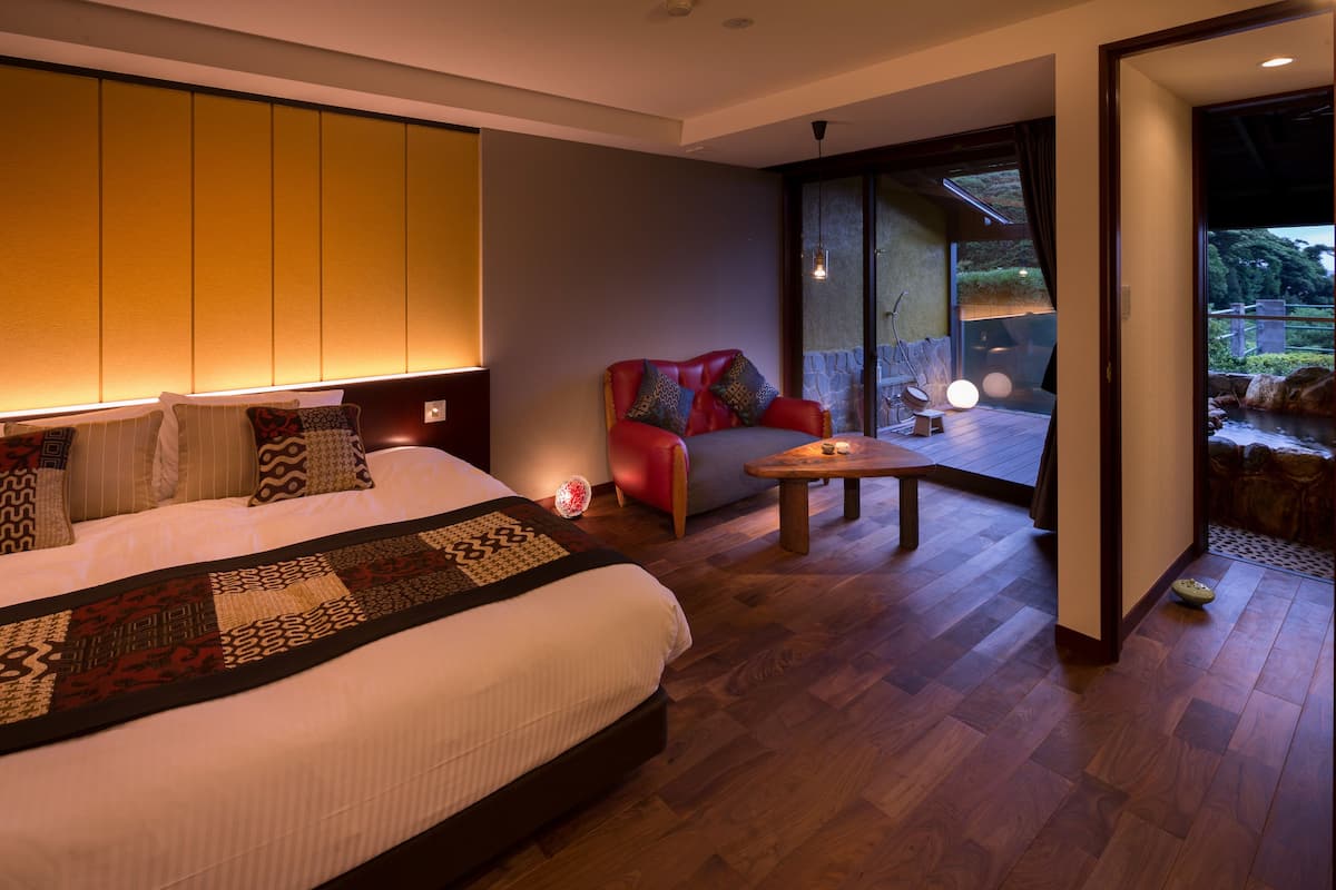 A modern hotel room with a large bed, a seating area with a red chair, and a view of a garden through a glass door.