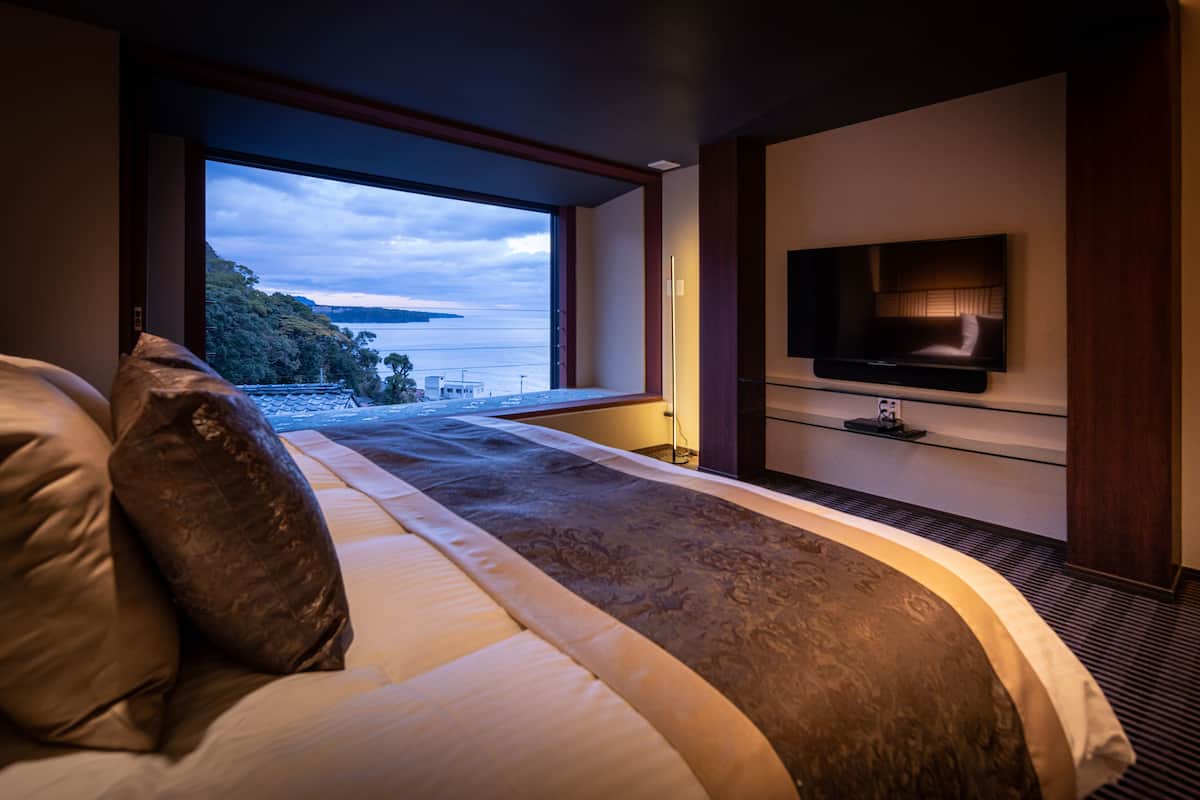 A modern bedroom with a large window, a flat-screen TV mounted on the wall, and a bed with a headboard.