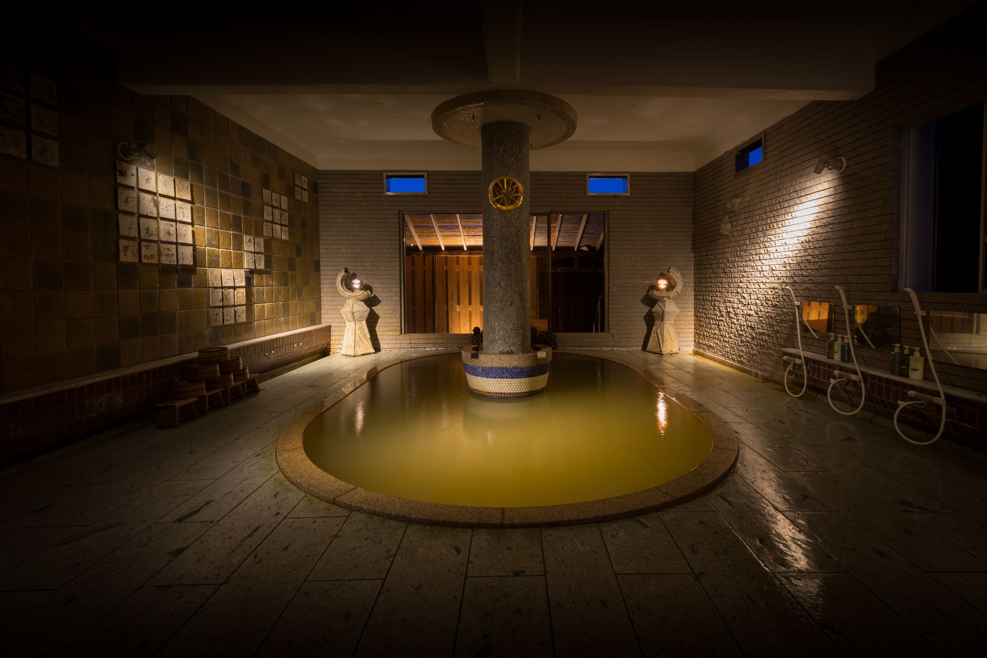 Zona de piscina interior con poca luz, una columna central, paredes con azulejos y suelo de madera.