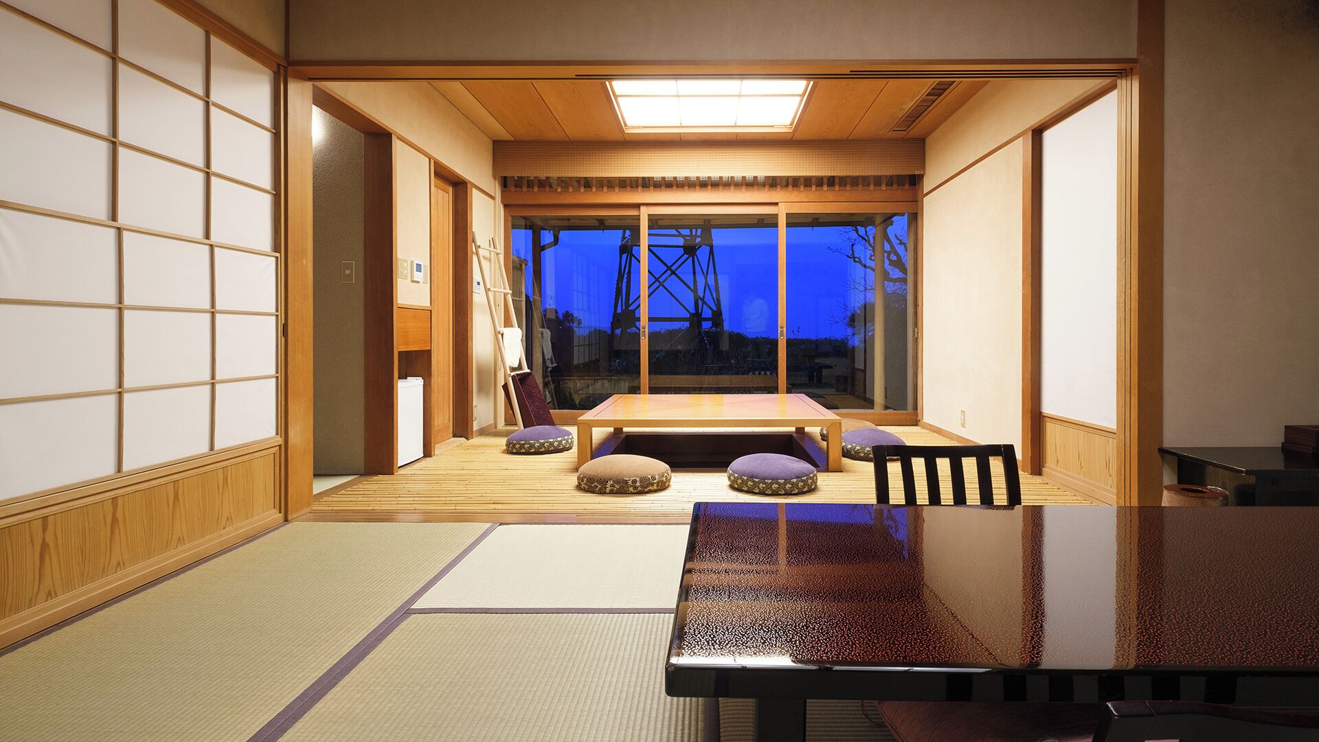 Ett traditionellt japanskt rum med tatamimattor på golvet, ett lågt bord, kuddar, skjutdörrar och utsikt över omgivningen.