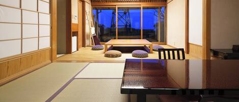 A traditional Japanese room with tatami flooring, a low table, cushions, sliding doors, and a view of the outside.