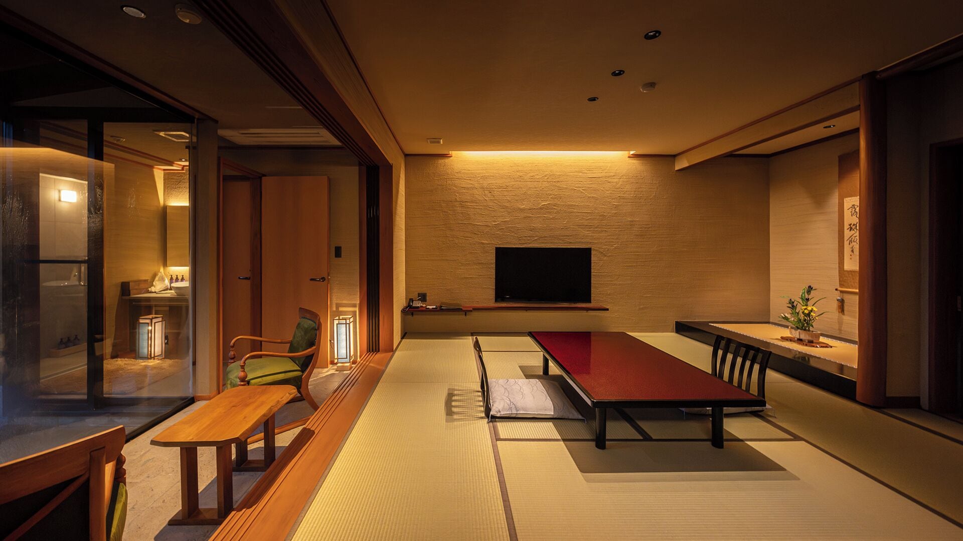 A traditional Japanese-style room with tatami flooring, a low table with a red mat, a wooden chair, a television mounted on the wall, and a sliding door leading to a bathroom.