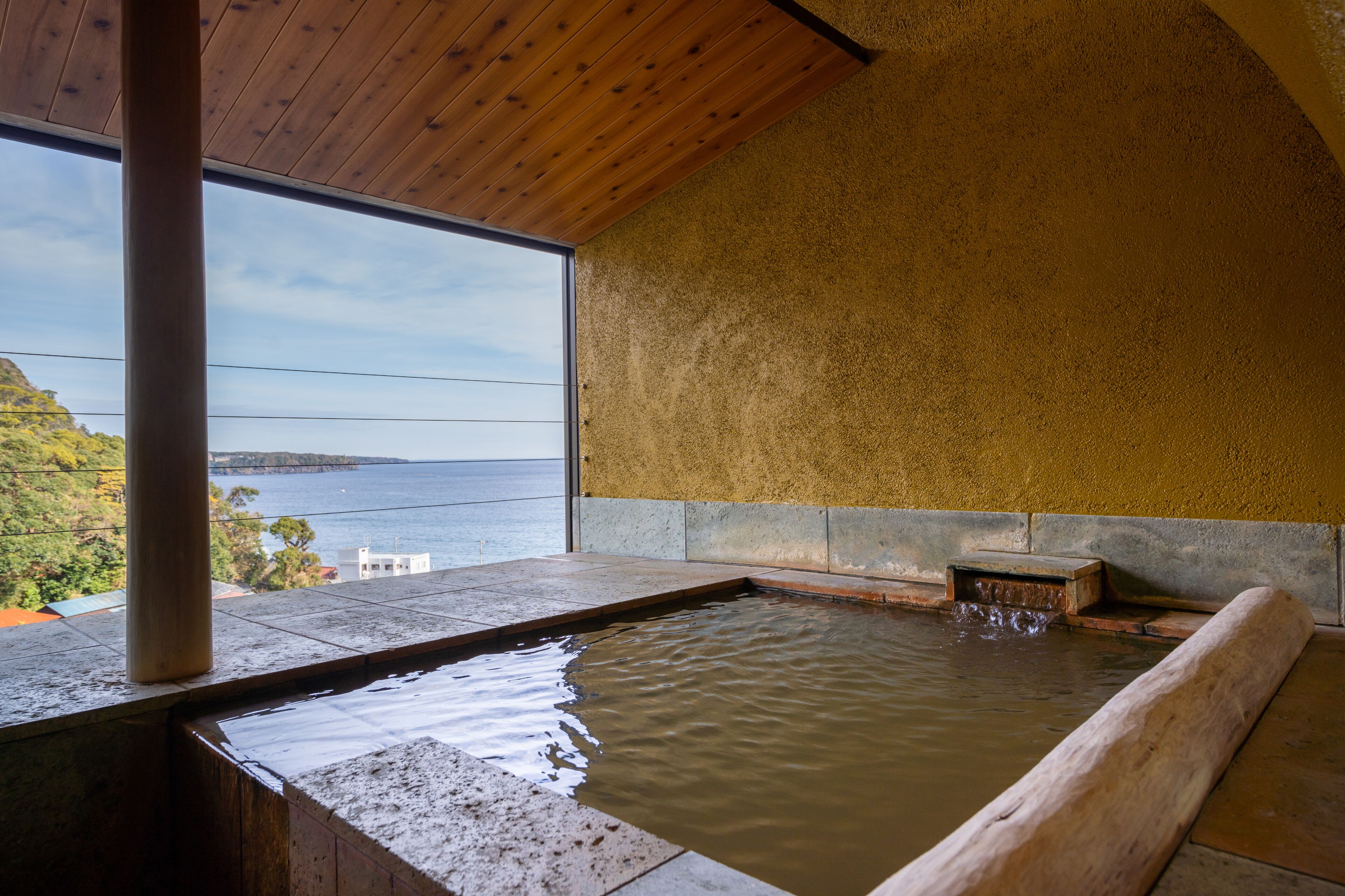 A hot spring bath with a wooden bench, a stone wall, and a view of the sea.