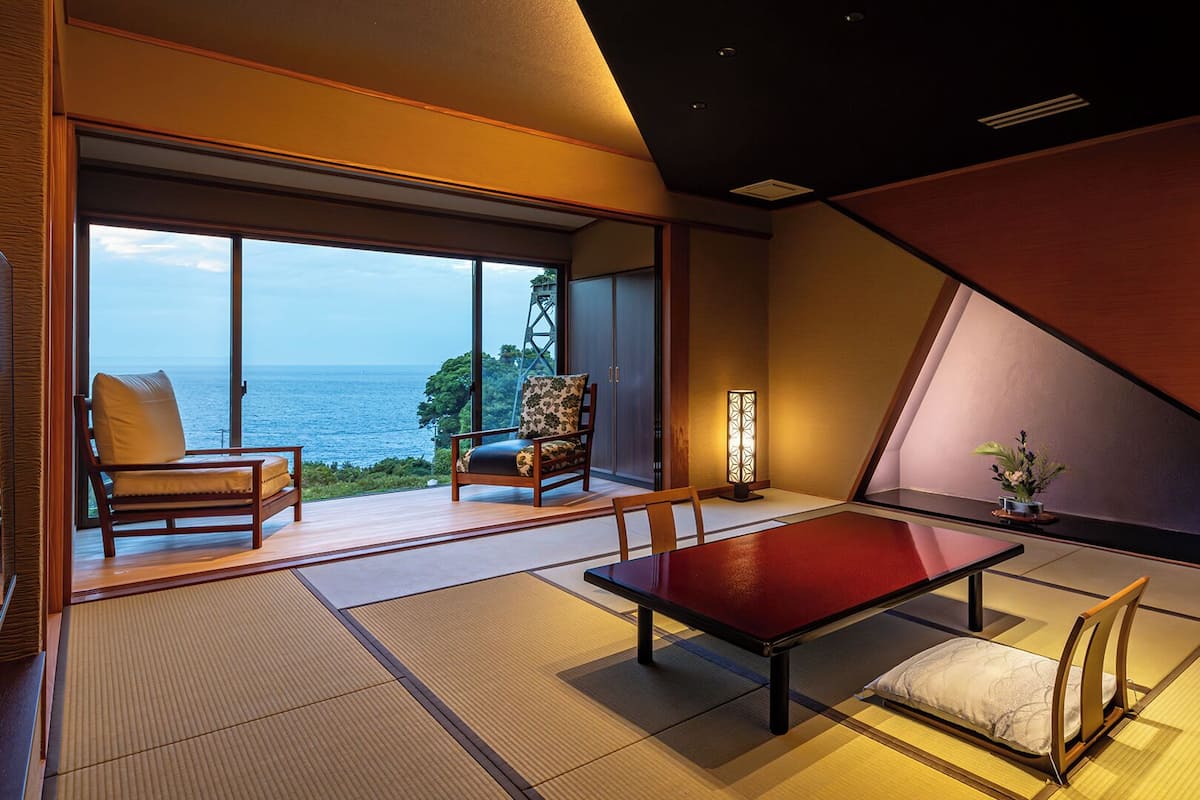 A modern interior with a large sliding glass door leading to a balcony with a view of the sea. Inside, there is a red table with a bench, a chair, and a floor lamp.