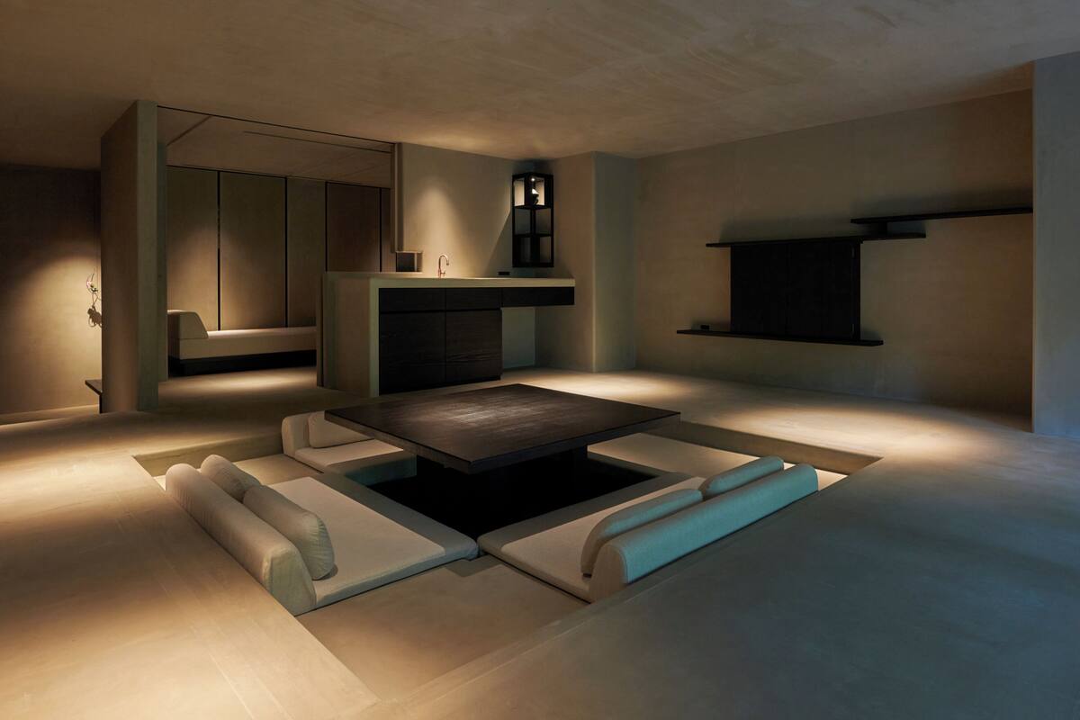 A modern, minimalist living room with a central coffee table, built-in seating, a kitchen area with a sink and cabinets, and a flat-screen TV mounted on the wall.