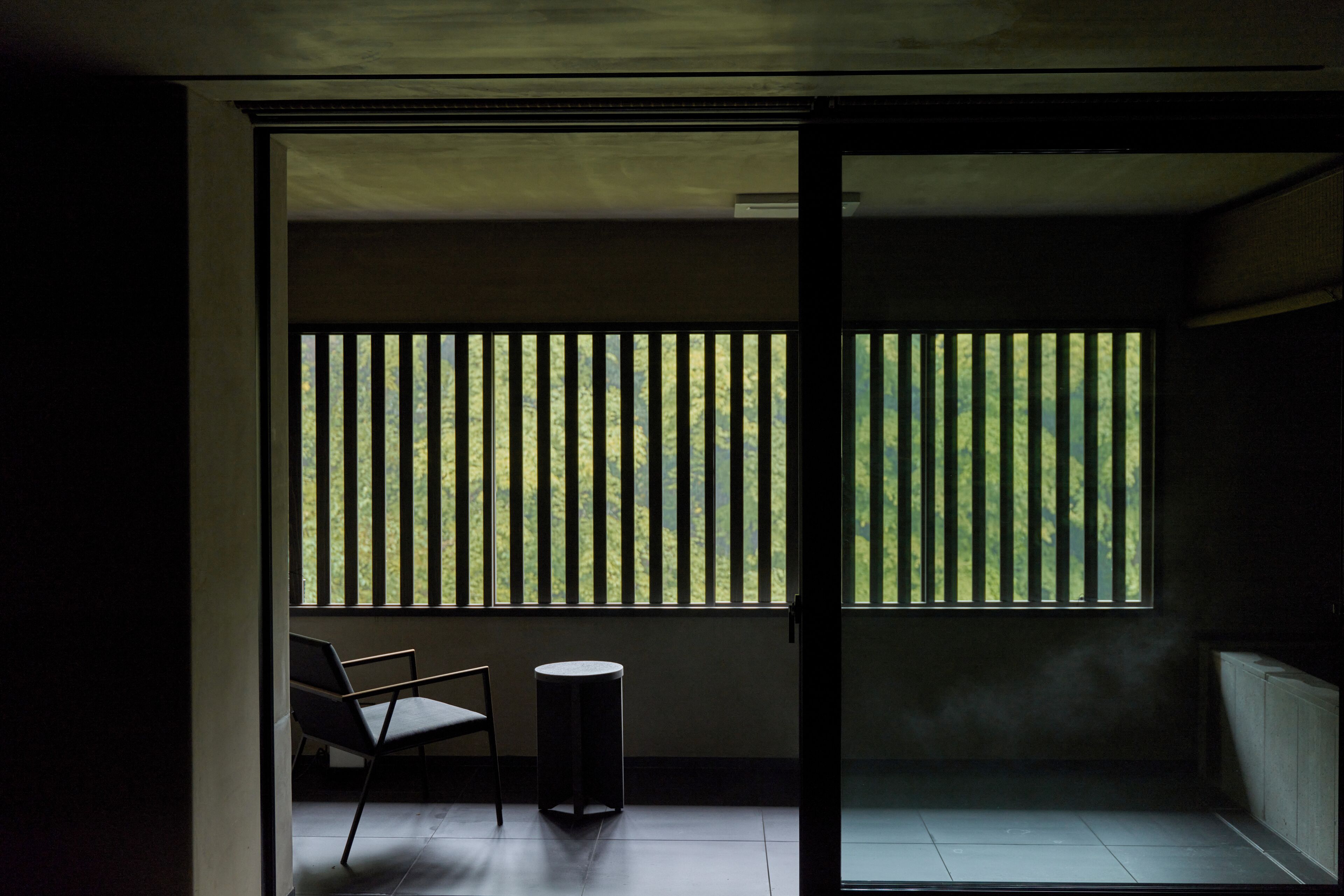 Una habitación con vista al exterior, visible a través de una ventana con persianas verticales, una silla y una pequeña mesa redonda.