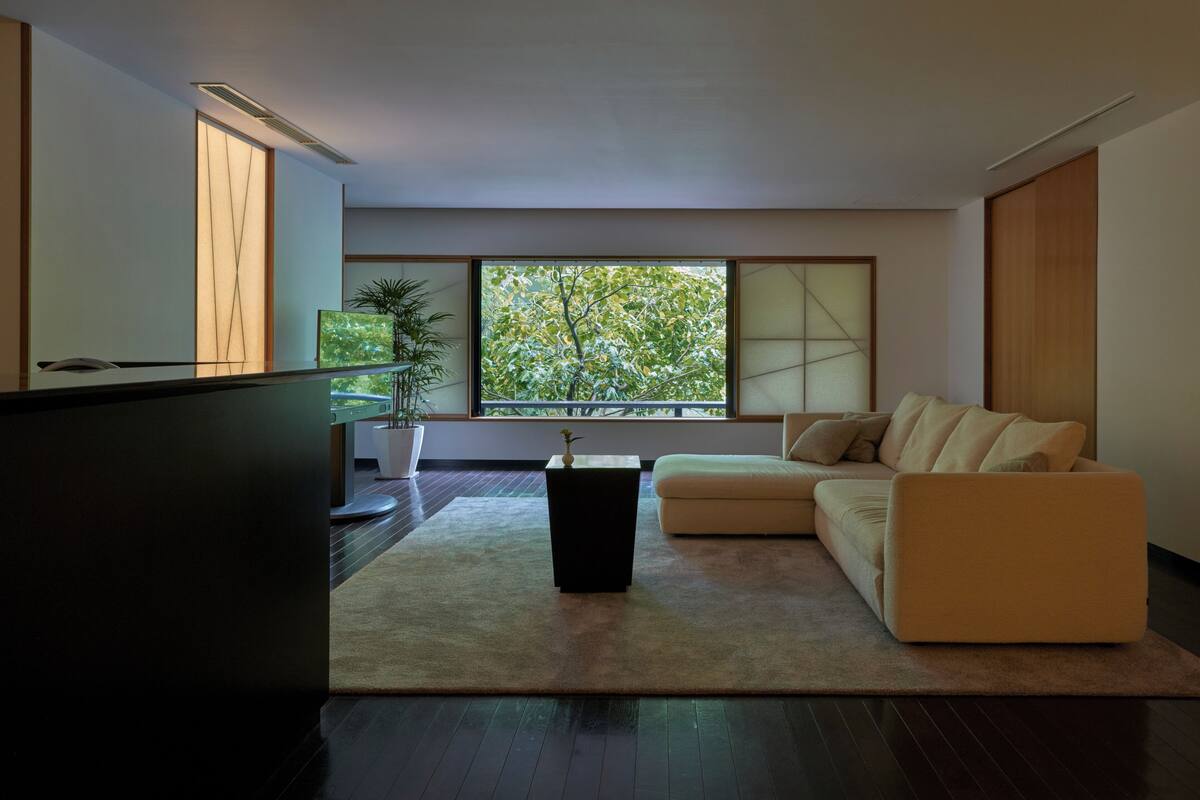 A modern living room with a sofa, a coffee table, and a TV. There is a large window with a view of trees.
