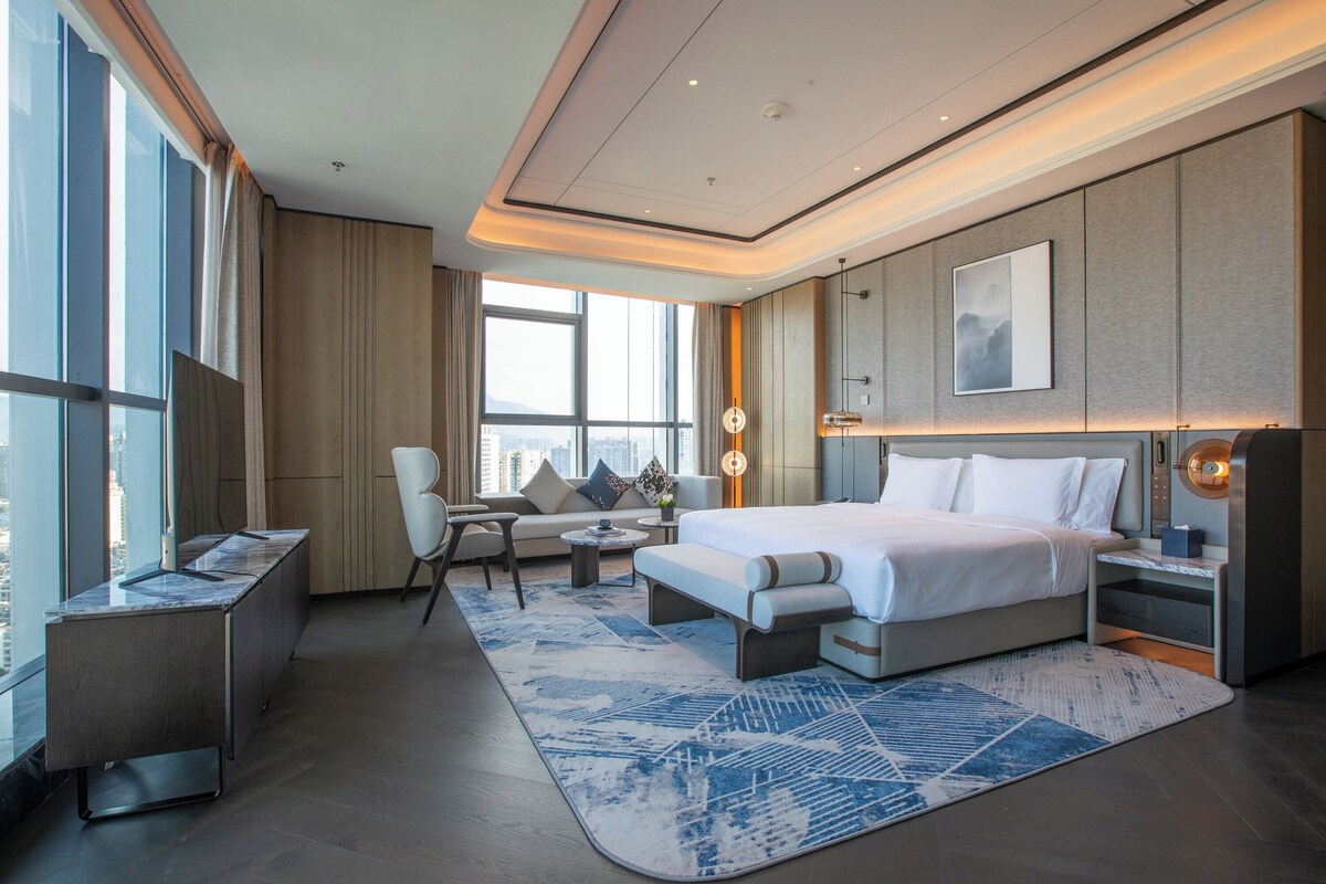 A modern hotel room with a large bed, a desk, a chair, and a sofa. There is a large window providing a city view.